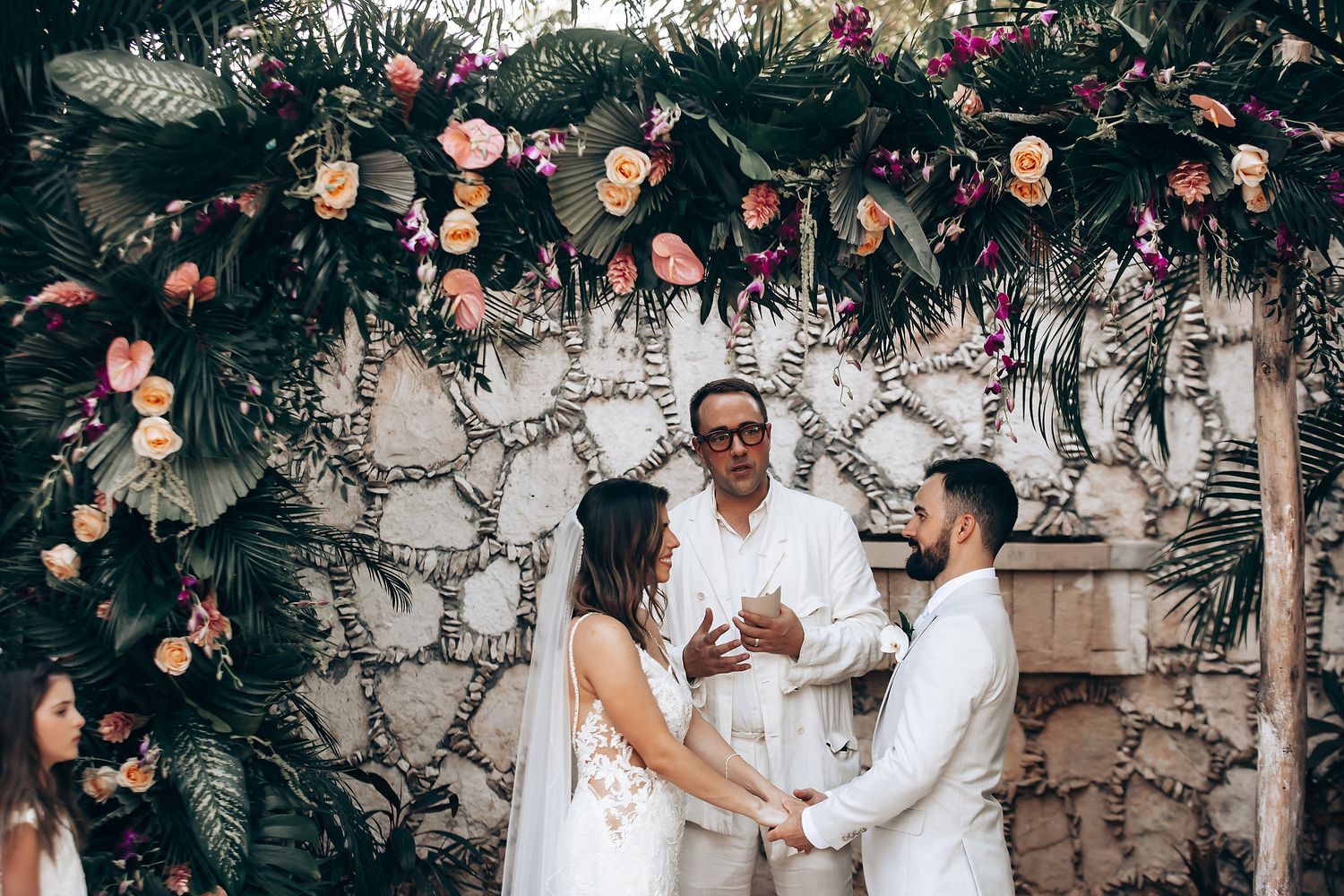 Intimate wedding ceremony moment under floral arch with stone wall backdrop.