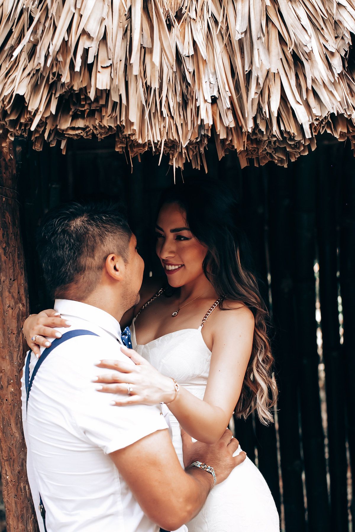 A romantic couple embraces under a thatched roof structure in matching white outfits.