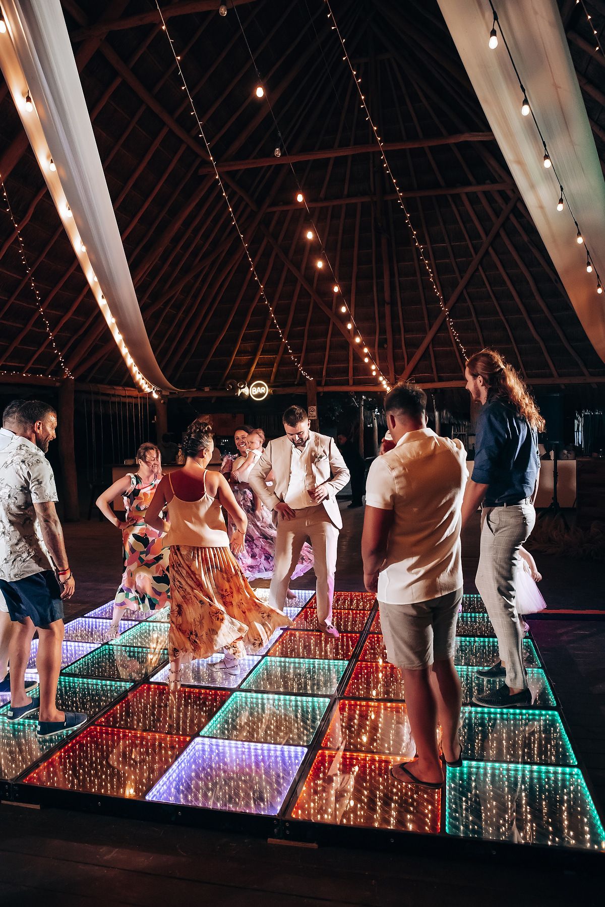 Party guests dancing on a colorful LED-lit dance floor under string lights in a barn reception venue.