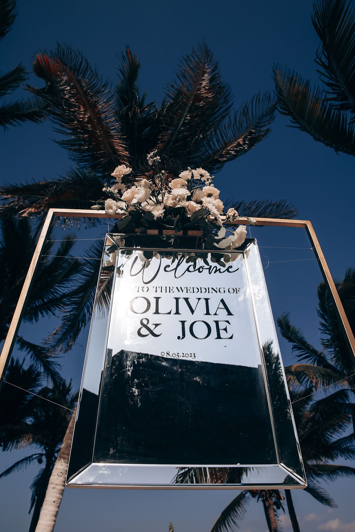 Wedding welcome sign displayed between palm trees against a blue sky at dusk with white floral decorations.