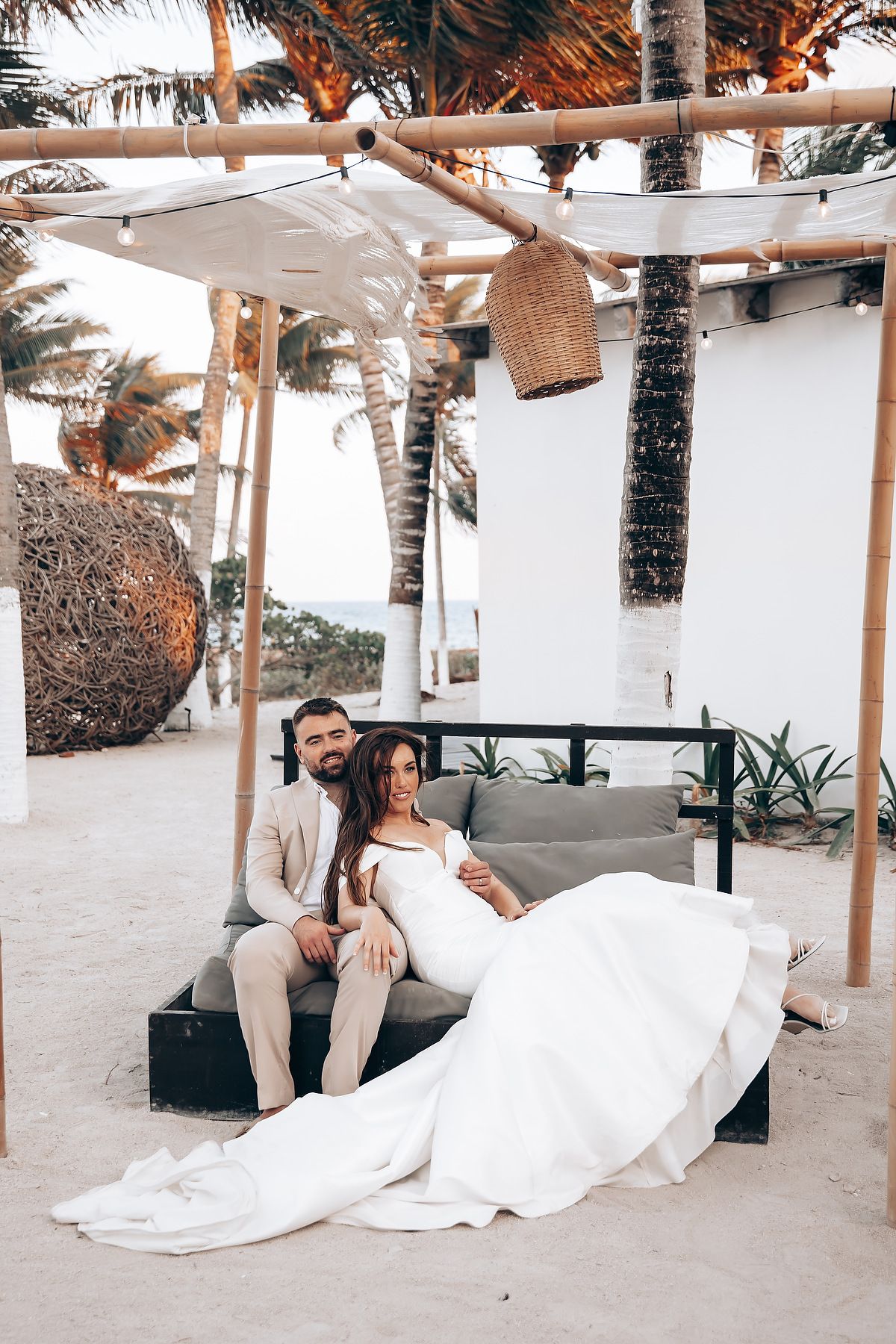 A couple relaxes together on white lounge chairs under palm trees at a tropical beach resort setting.