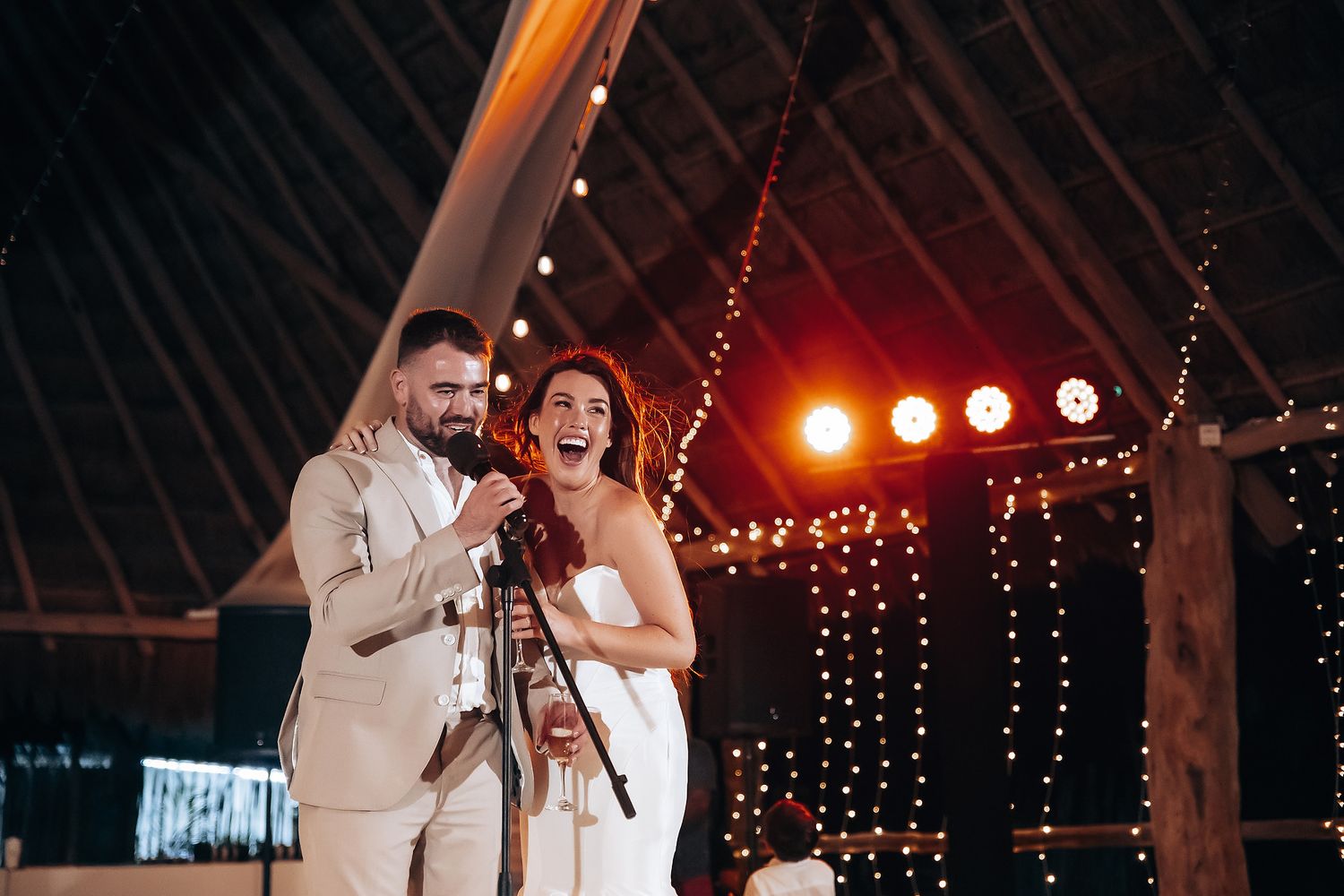 Wedding couple dances together beneath string lights and red stage lighting in a rustic venue.