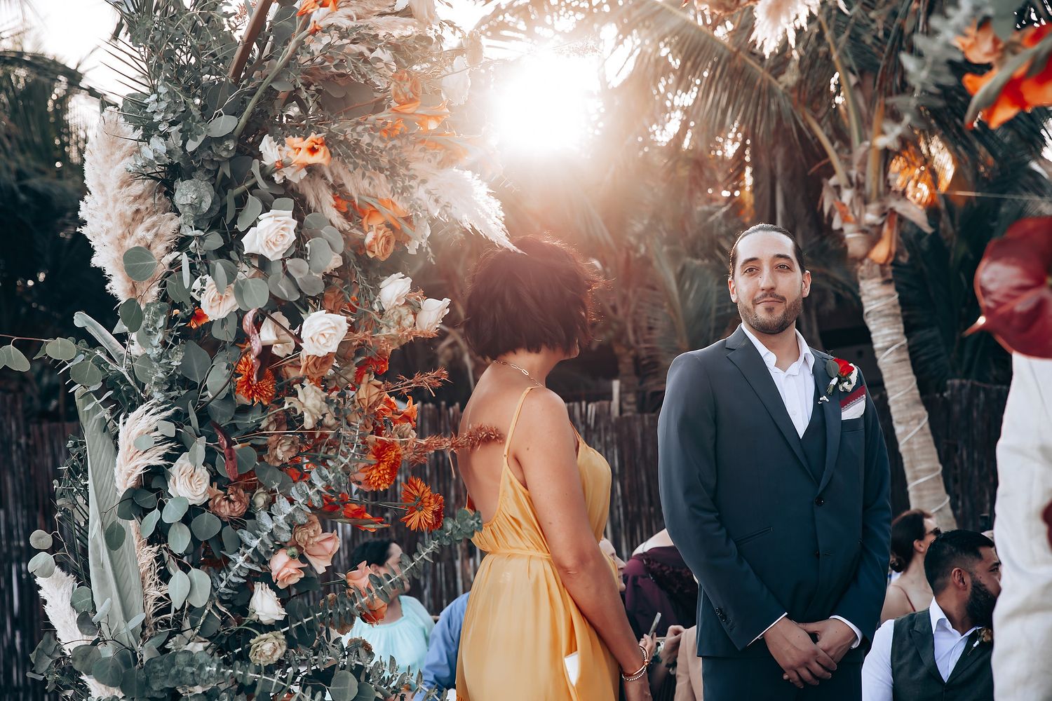 Outdoor wedding ceremony with floral arch decorations and palm trees in natural sunlight.