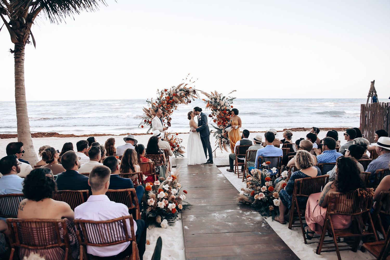 Oceanfront wedding ceremony setup with guests seated under palm trees.
