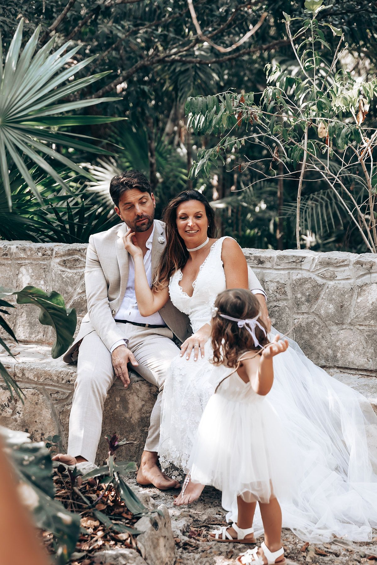 A romantic wedding photoshoot in a tropical garden setting with palm fronds and stone walls.