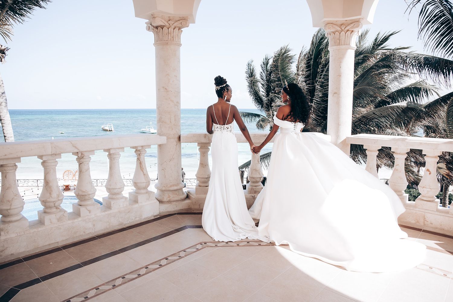 Two brides in flowing white gowns pose against a scenic oceanfront balcony with palm trees.
