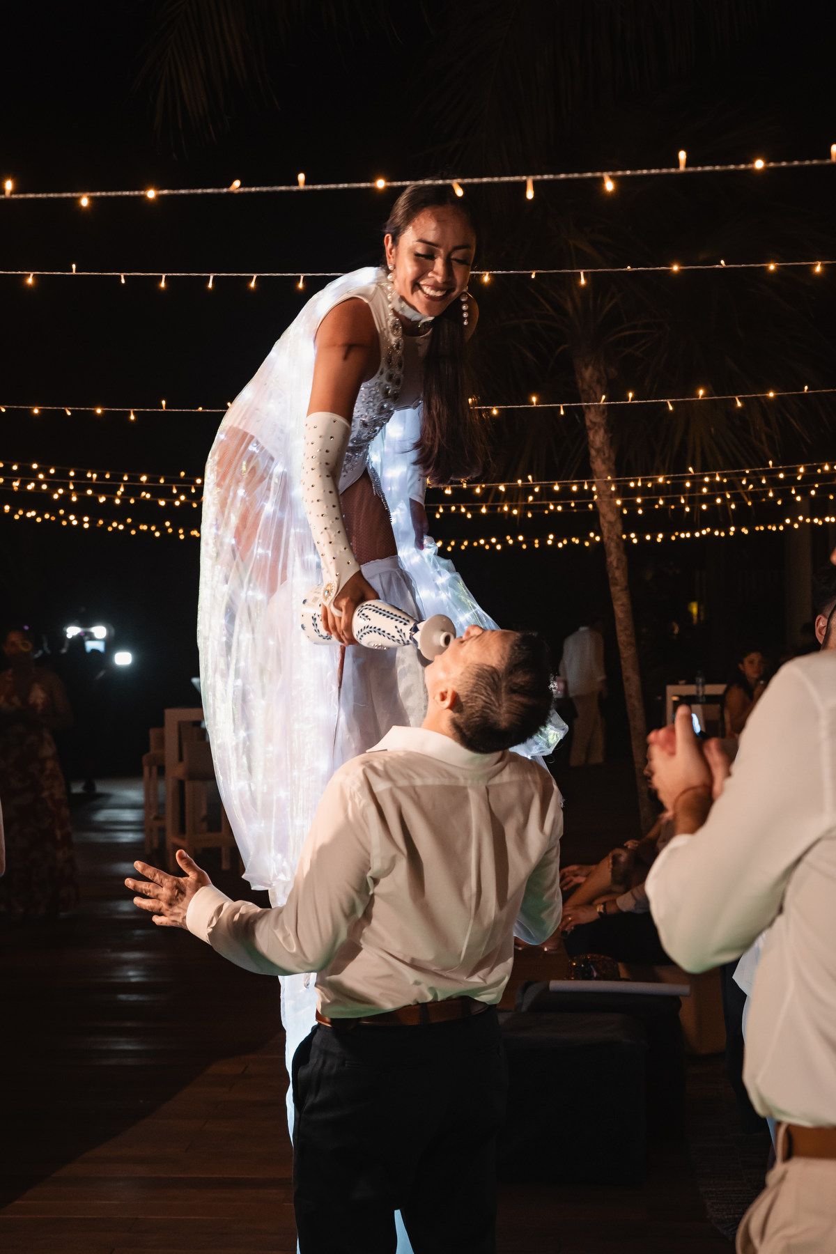 Groom lifts bride in sparkling reception dress on outdoor dance floor under string lights.