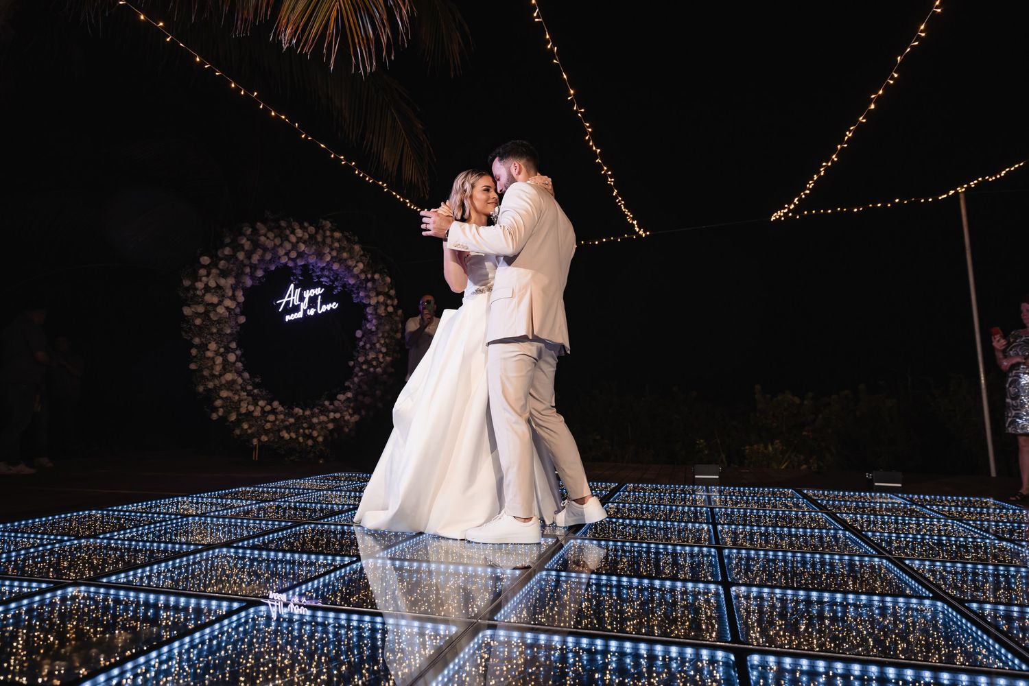 Bride and groom share first dance on illuminated glass floor under string lights at night.