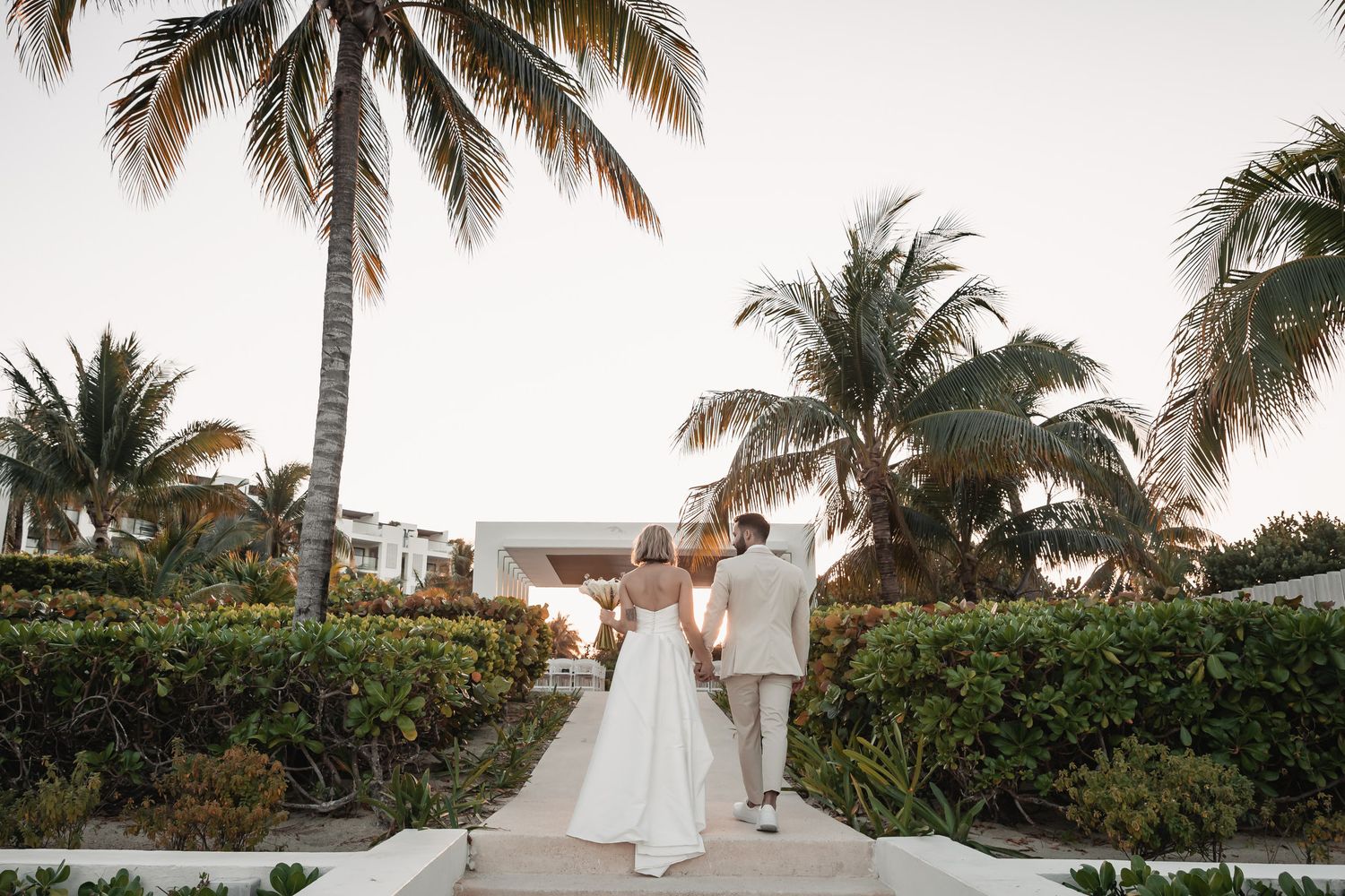 Bride and groom walking hand-in-hand through tropical palm tree lined resort path.