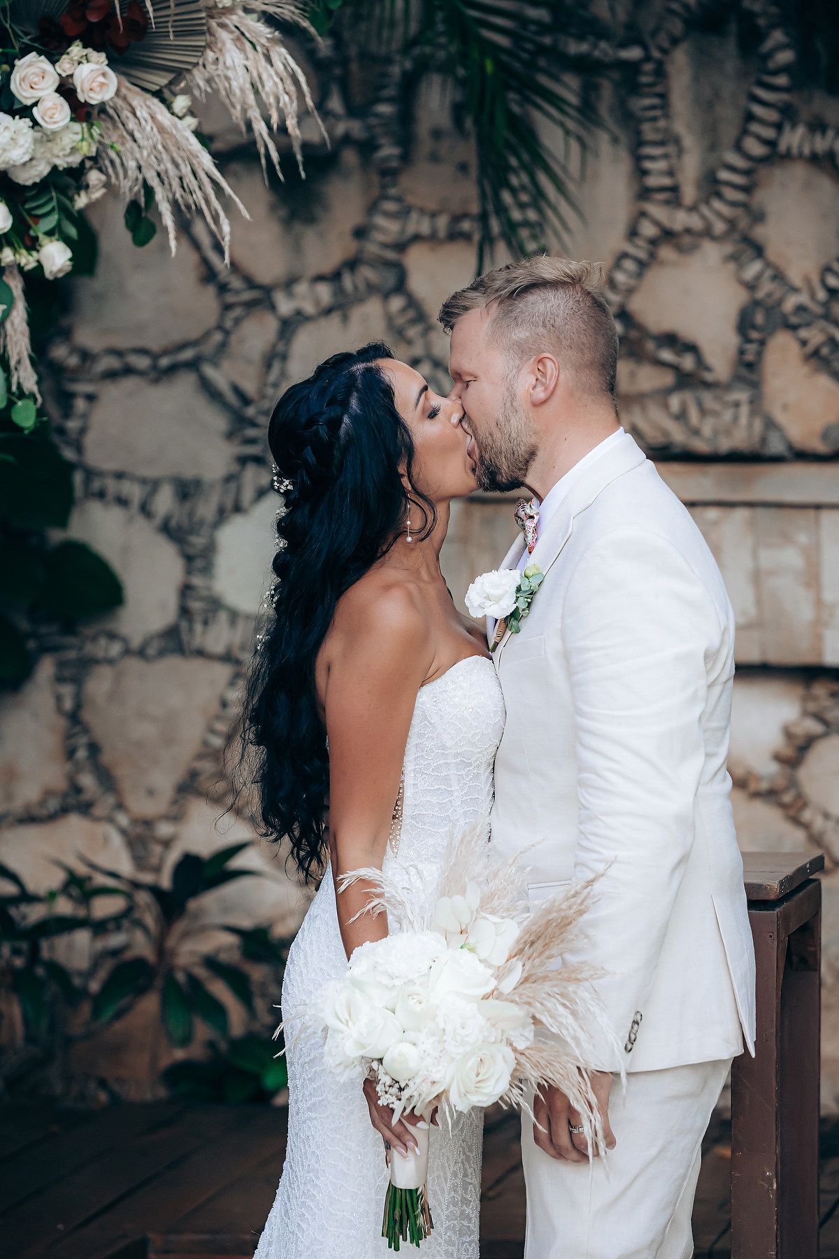 Romantic wedding couple in white attire sharing an intimate moment against a textured wall.
