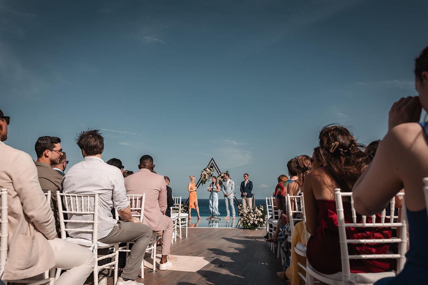Wedding guests seated in white chairs at outdoor oceanfront ceremony.