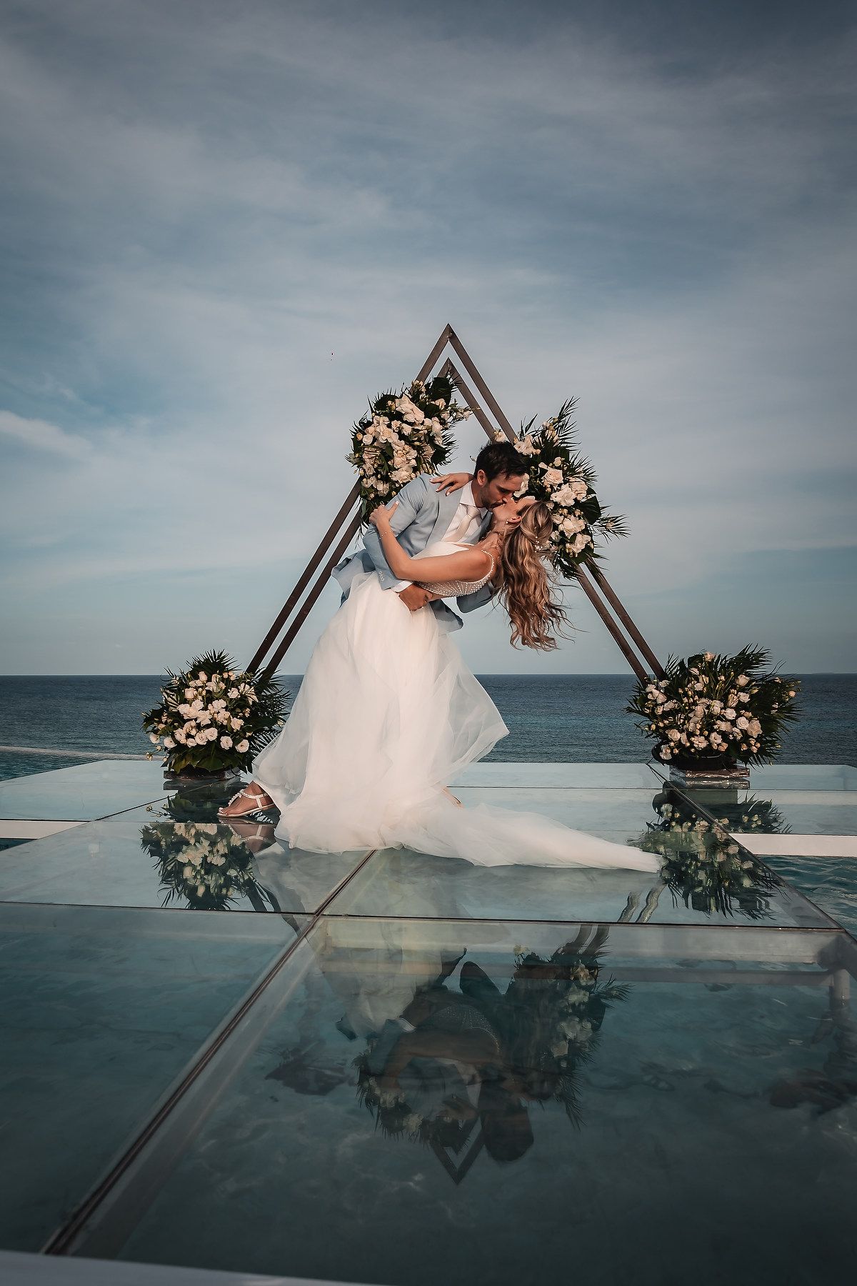 Wedding couple embracing under geometric arch with flowers and ocean view.