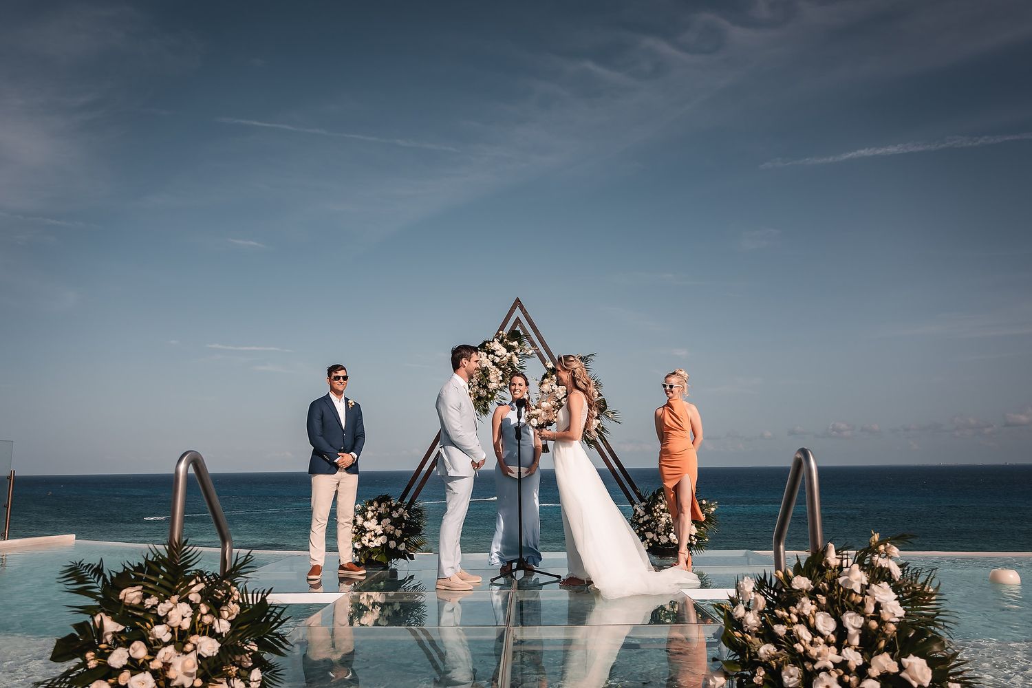 Beachfront wedding ceremony setup with geometric arch and floral arrangements against blue ocean backdrop.