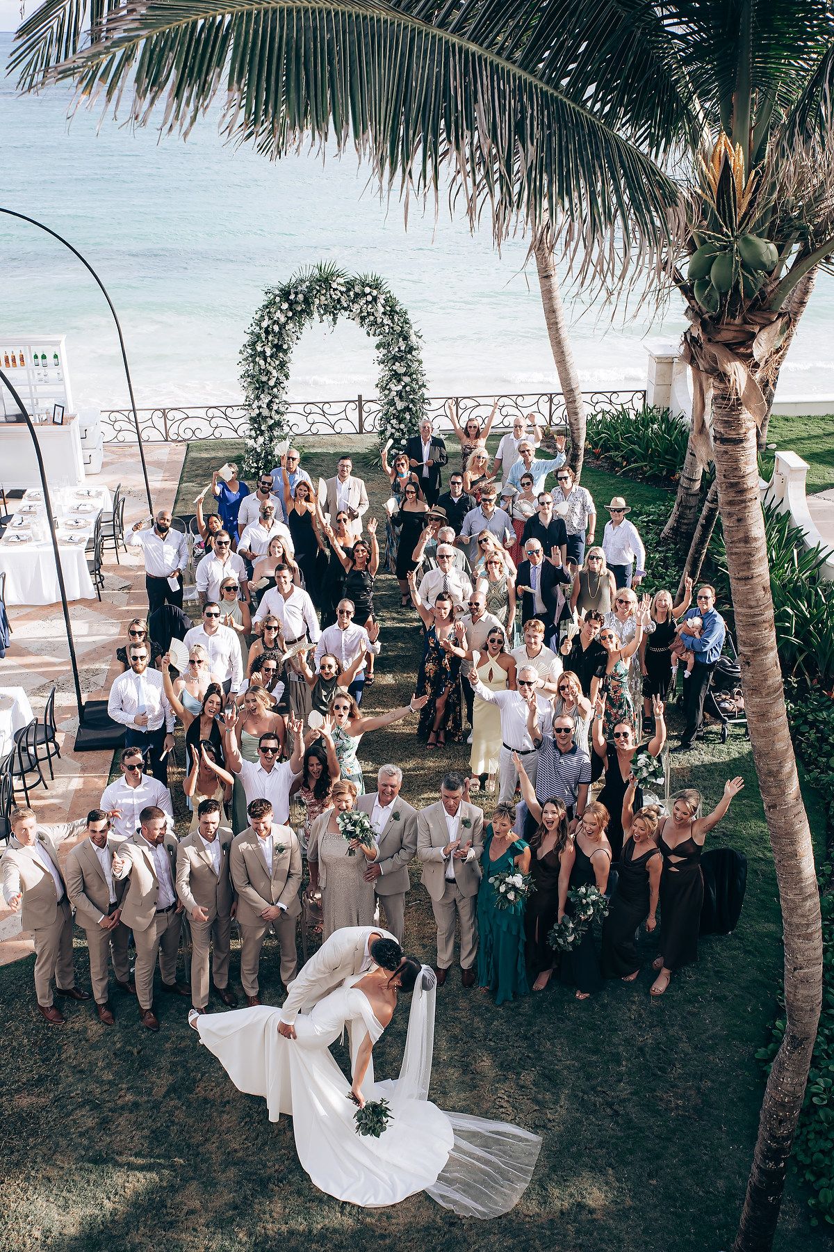 Beach wedding celebration with large group photo under palm trees by the ocean.