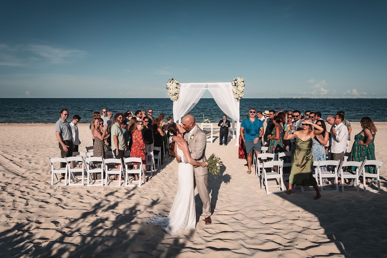 Wedding guests seated in white chairs on a sandy beach with an oceanfront ceremony arch.