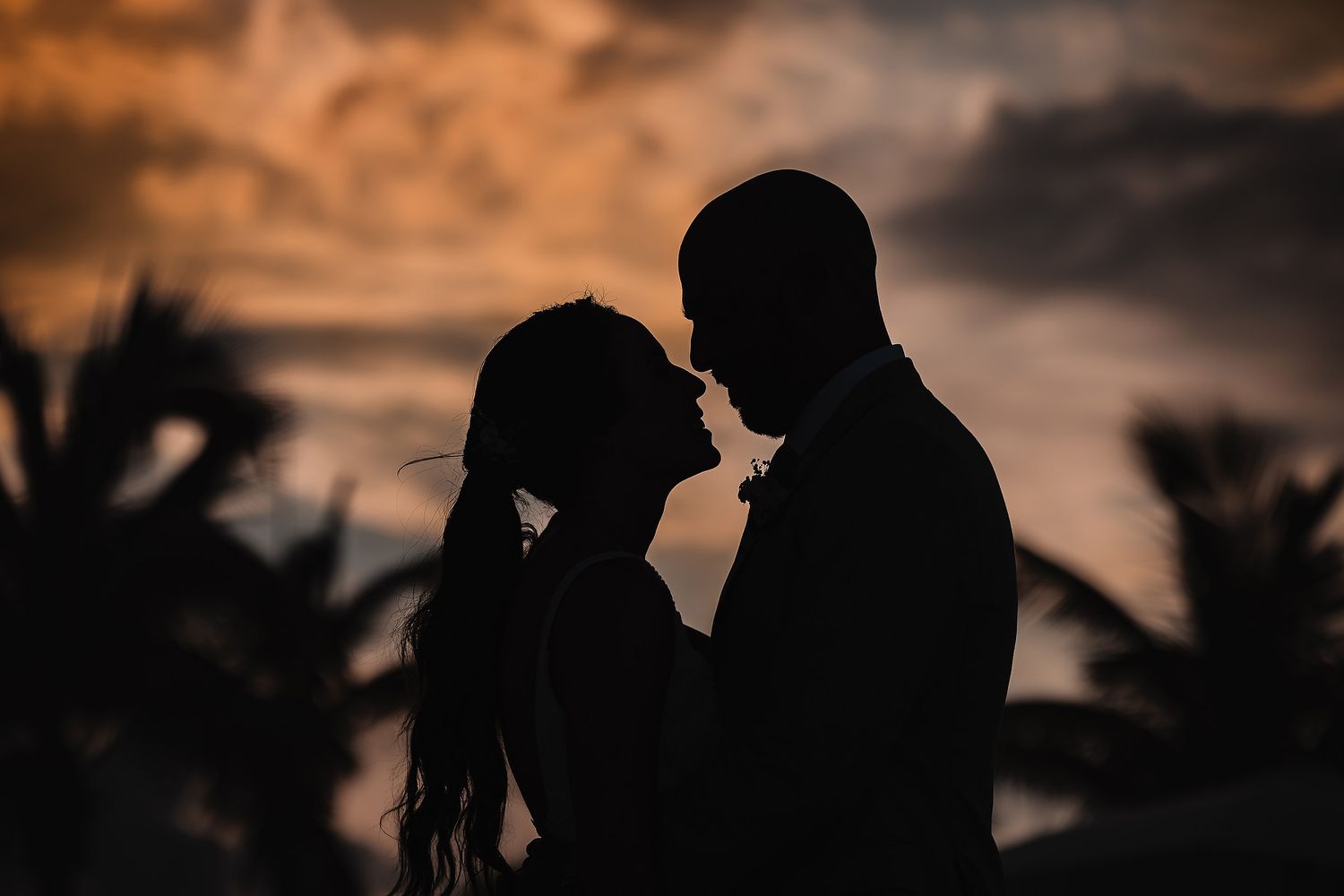 Palm trees create silhouettes against an orange sunset sky during a beach wedding.