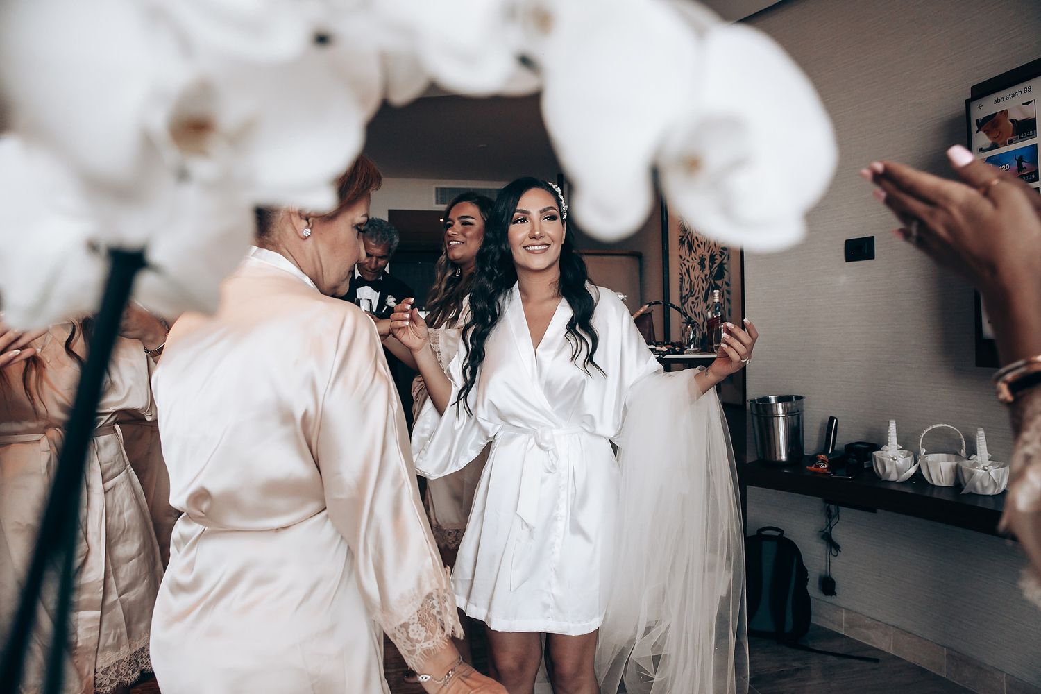 Bridesmaids in white robes helping with wedding preparations in a softly lit room.