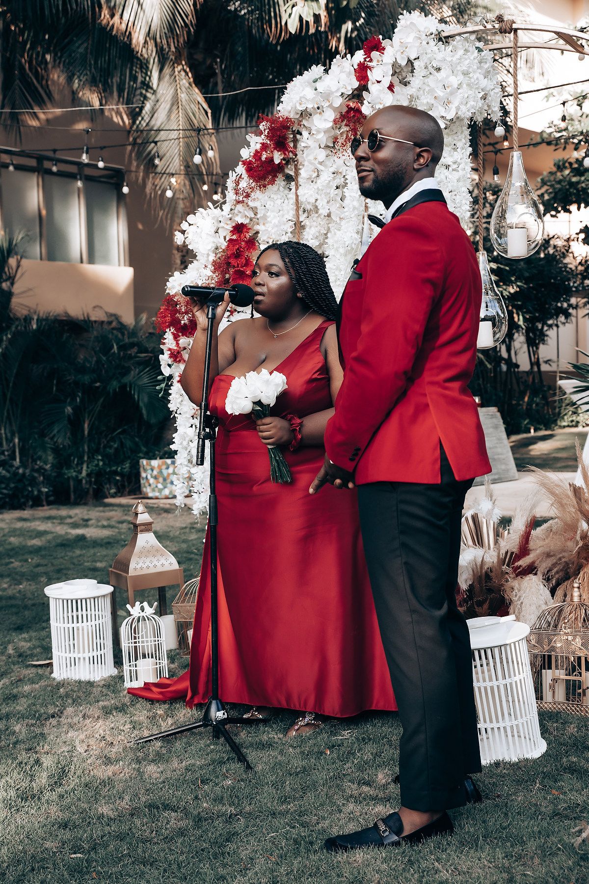 A couple in elegant red and black attire pose together in a garden setting with white decorative elements.