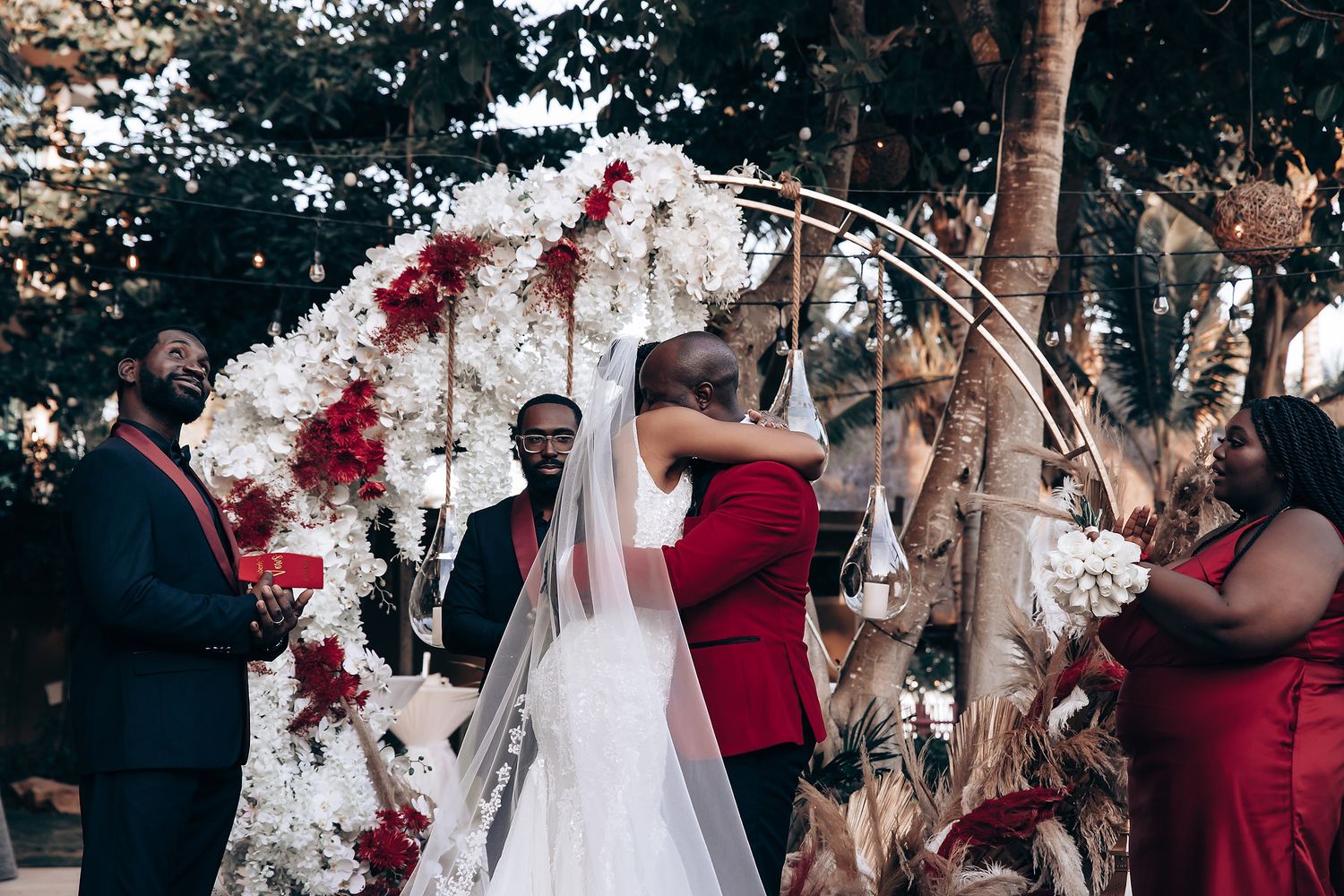 Outdoor wedding celebration with guests dancing beneath trees decorated with red and white floral accents.