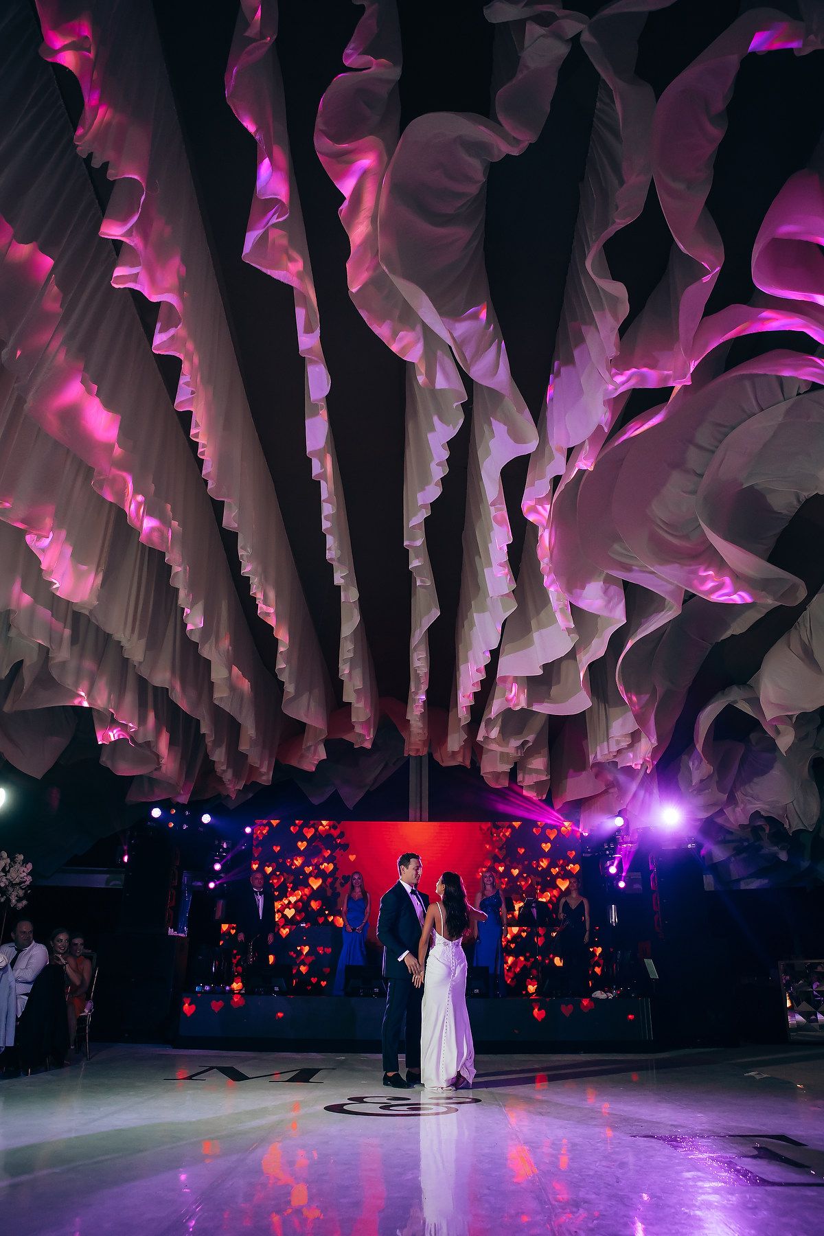 A couple dances under dramatic pink-lit fabric draping and heart-shaped decor during their wedding reception.