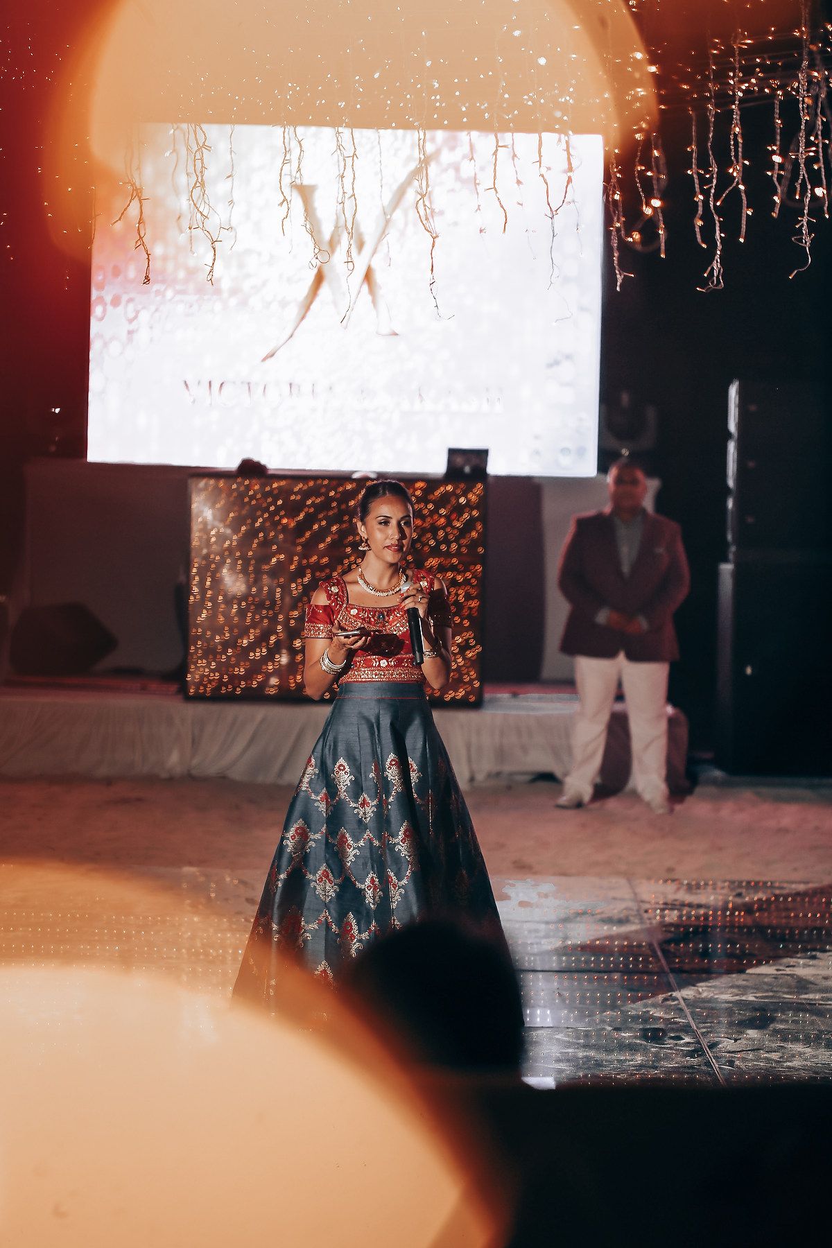 A model showcases an elegant navy blue evening gown with embroidered details on a dimly lit runway stage.