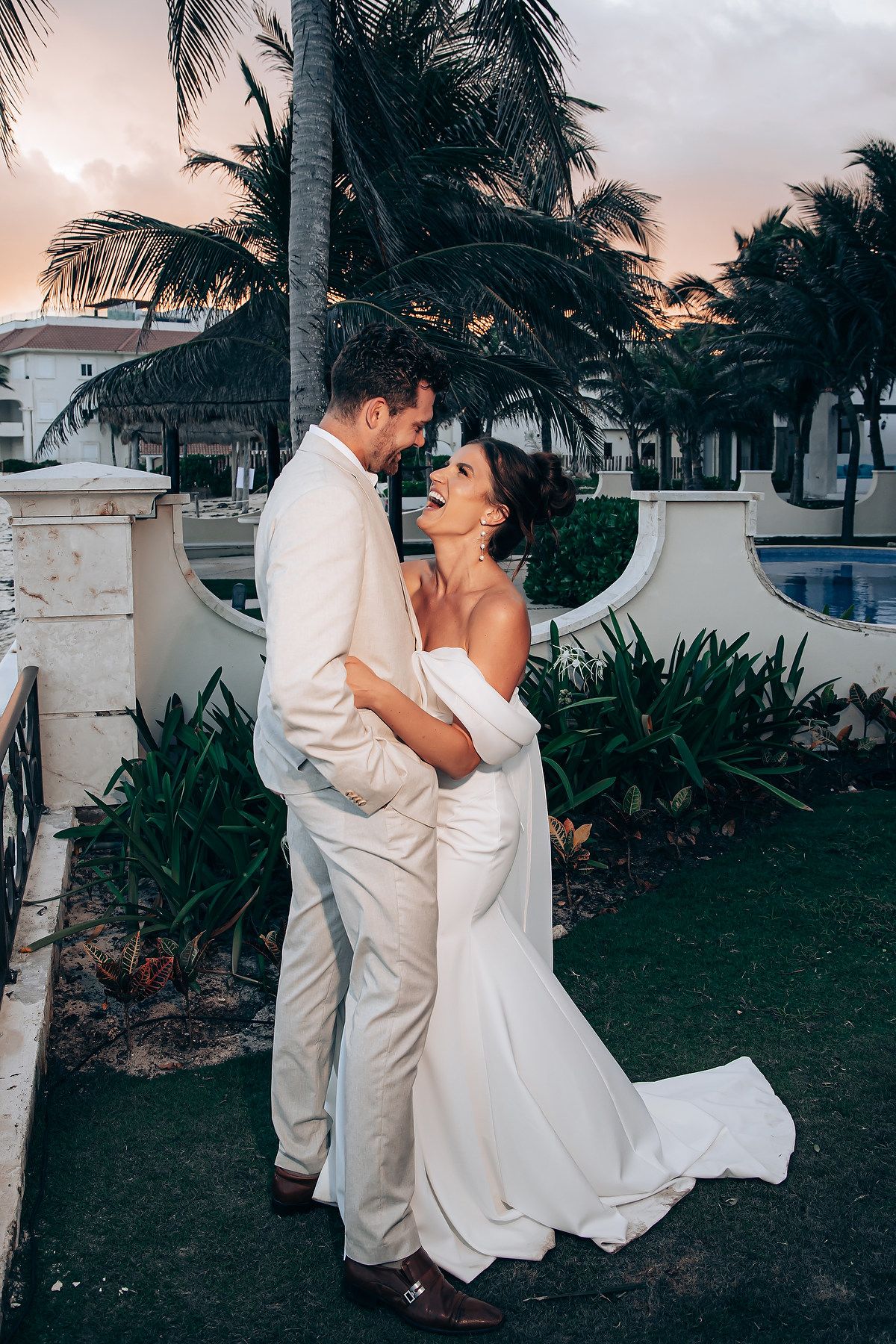 A romantic beach resort wedding couple embraces at sunset with palm trees and tropical landscaping in the background.