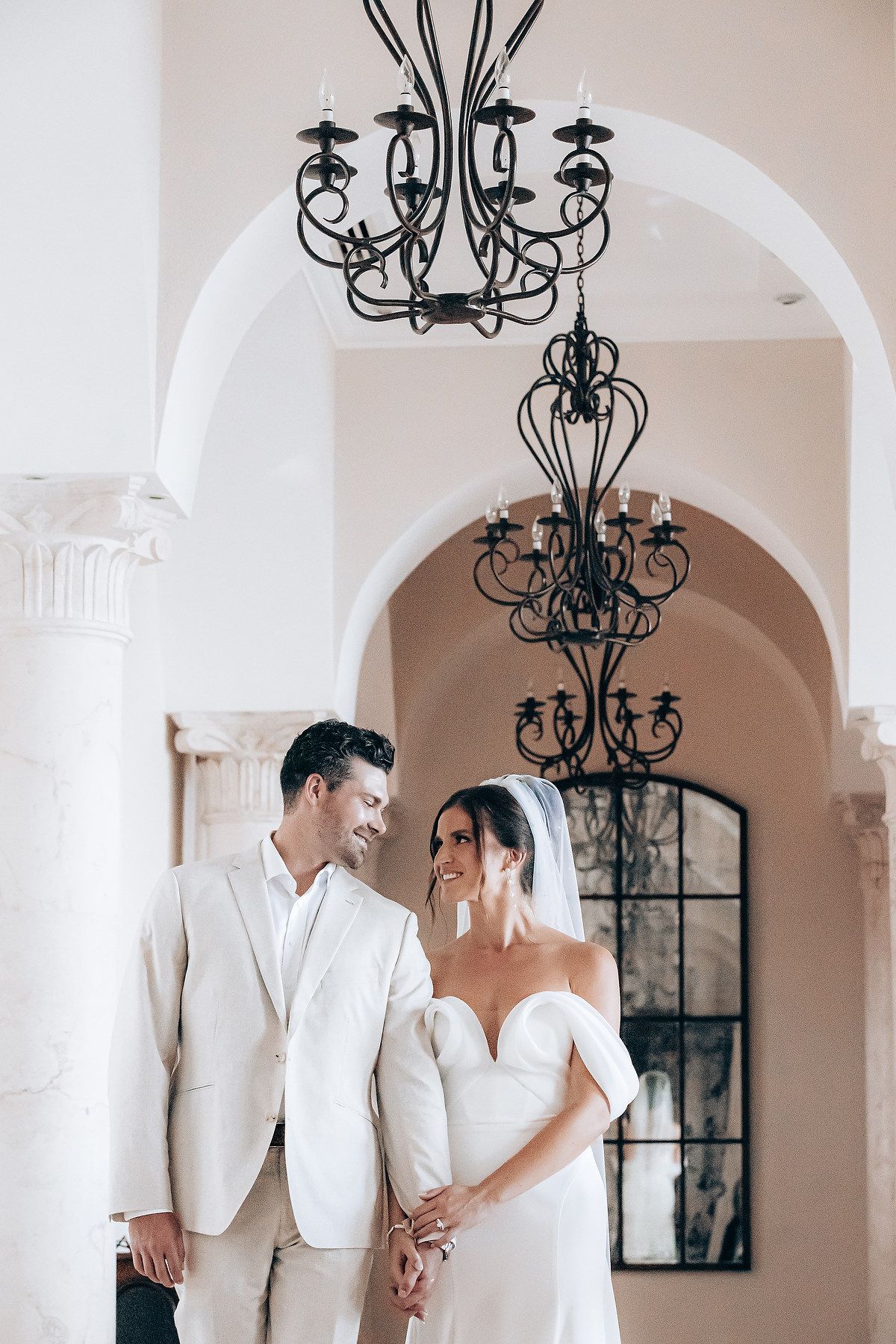 Elegant wedding couple in white attire standing beneath ornate black iron chandeliers in arched hallway.