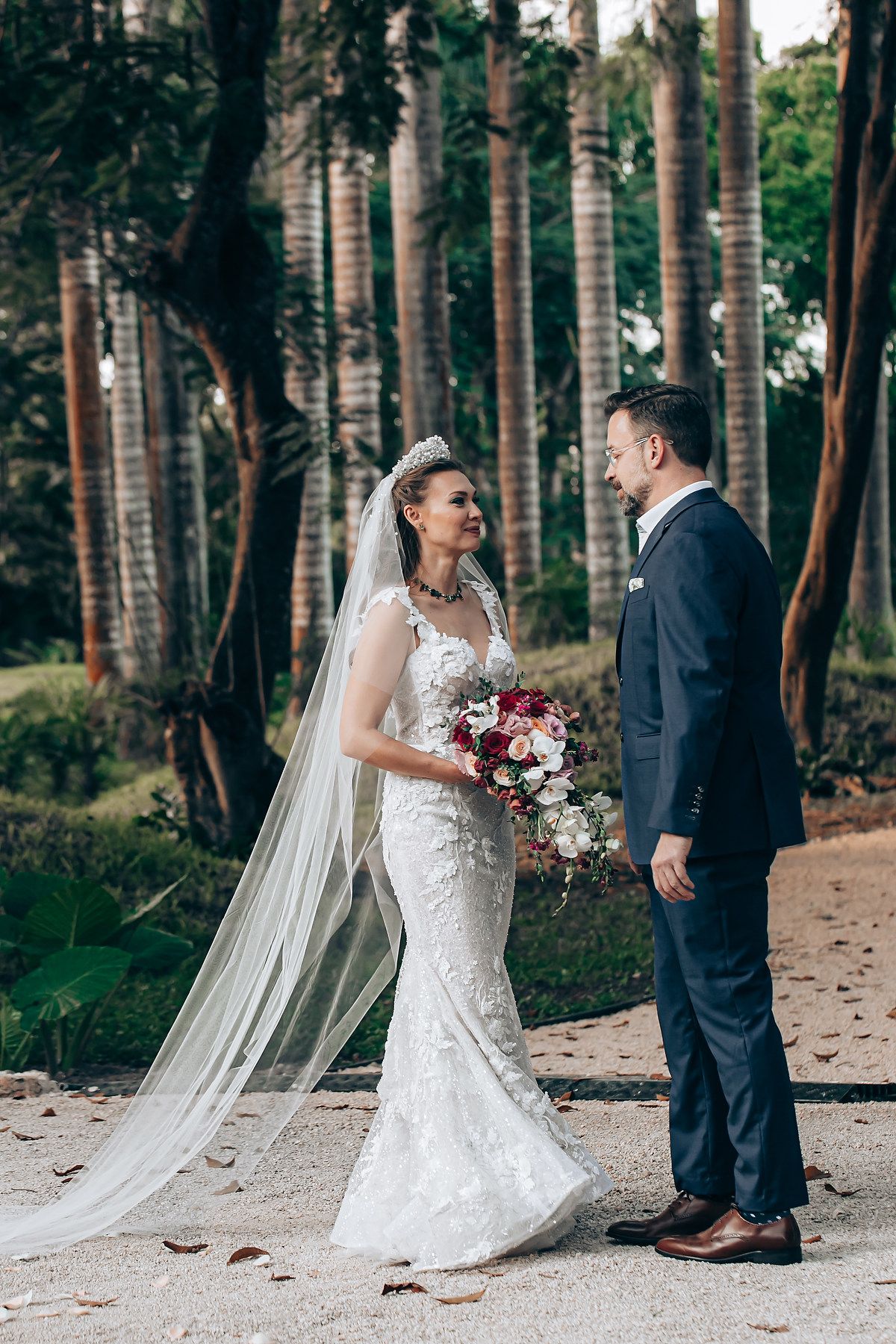 A romantic wedding couple stands together on a forest path with tall trees and a long flowing veil.