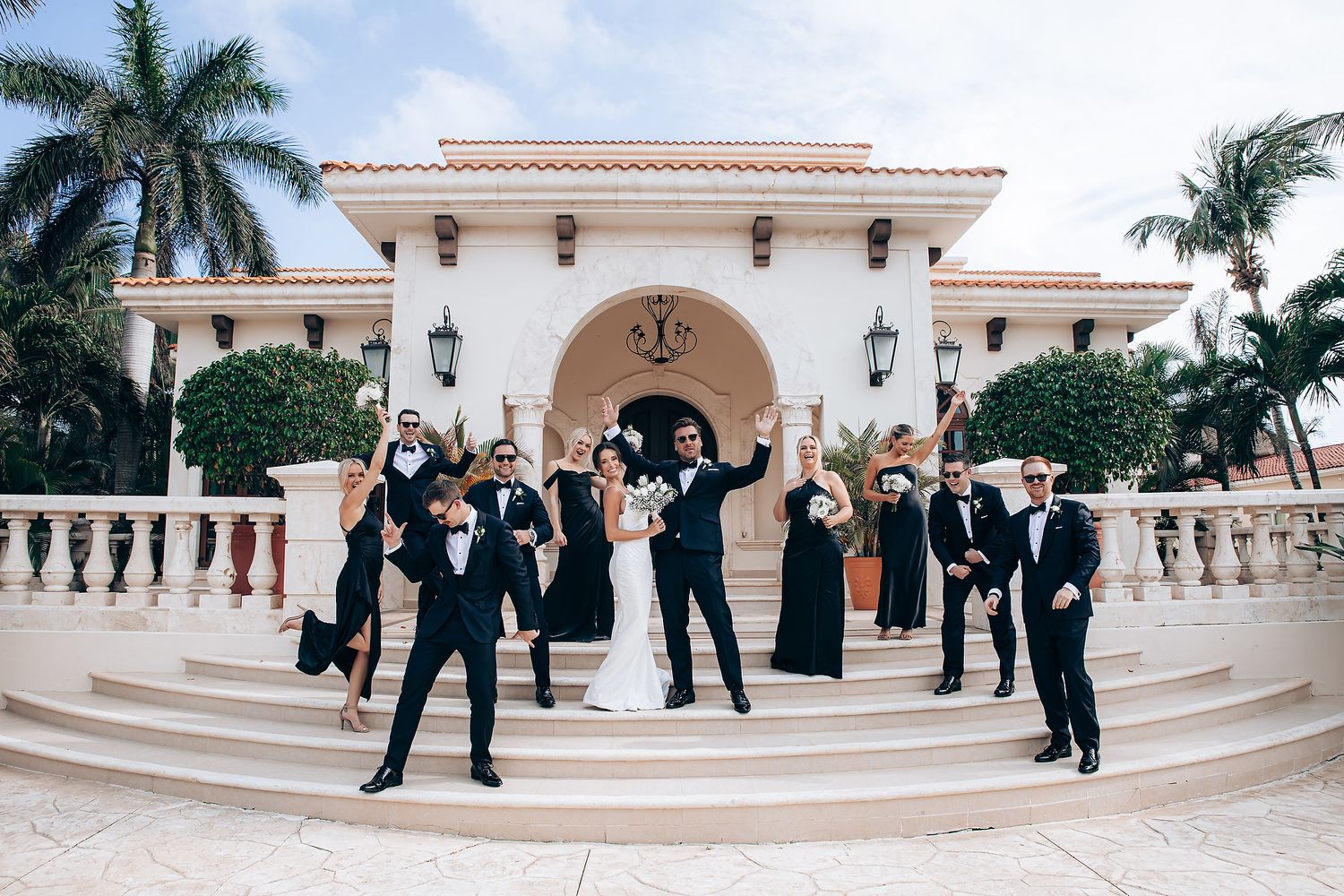 Wedding party in formal attire poses on grand white stone steps of Mediterranean-style mansion with palm trees.
