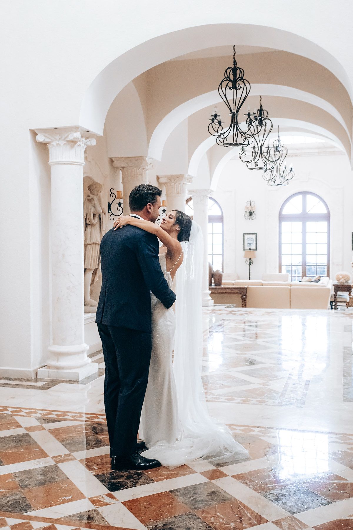 A romantic wedding moment in an elegant white marble hall with arched columns and chandeliers.
