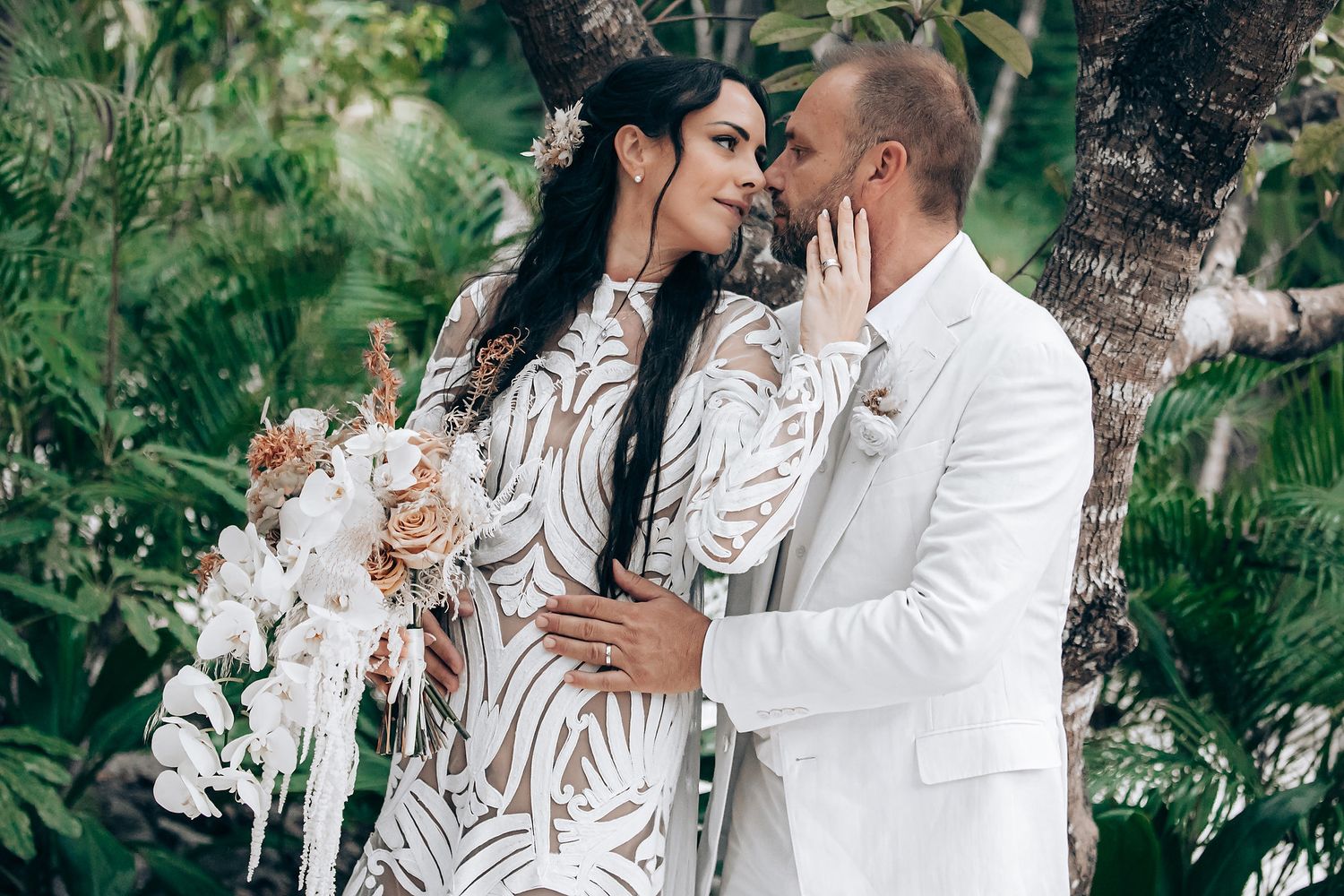 Romantic couple in white attire embracing outdoors surrounded by lush greenery.