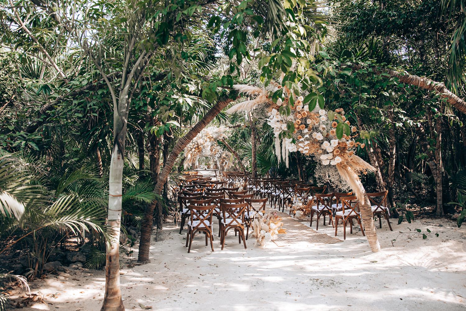 Rustic wooden chairs arranged for an outdoor wedding ceremony in a tropical forest setting with natural sunlight.