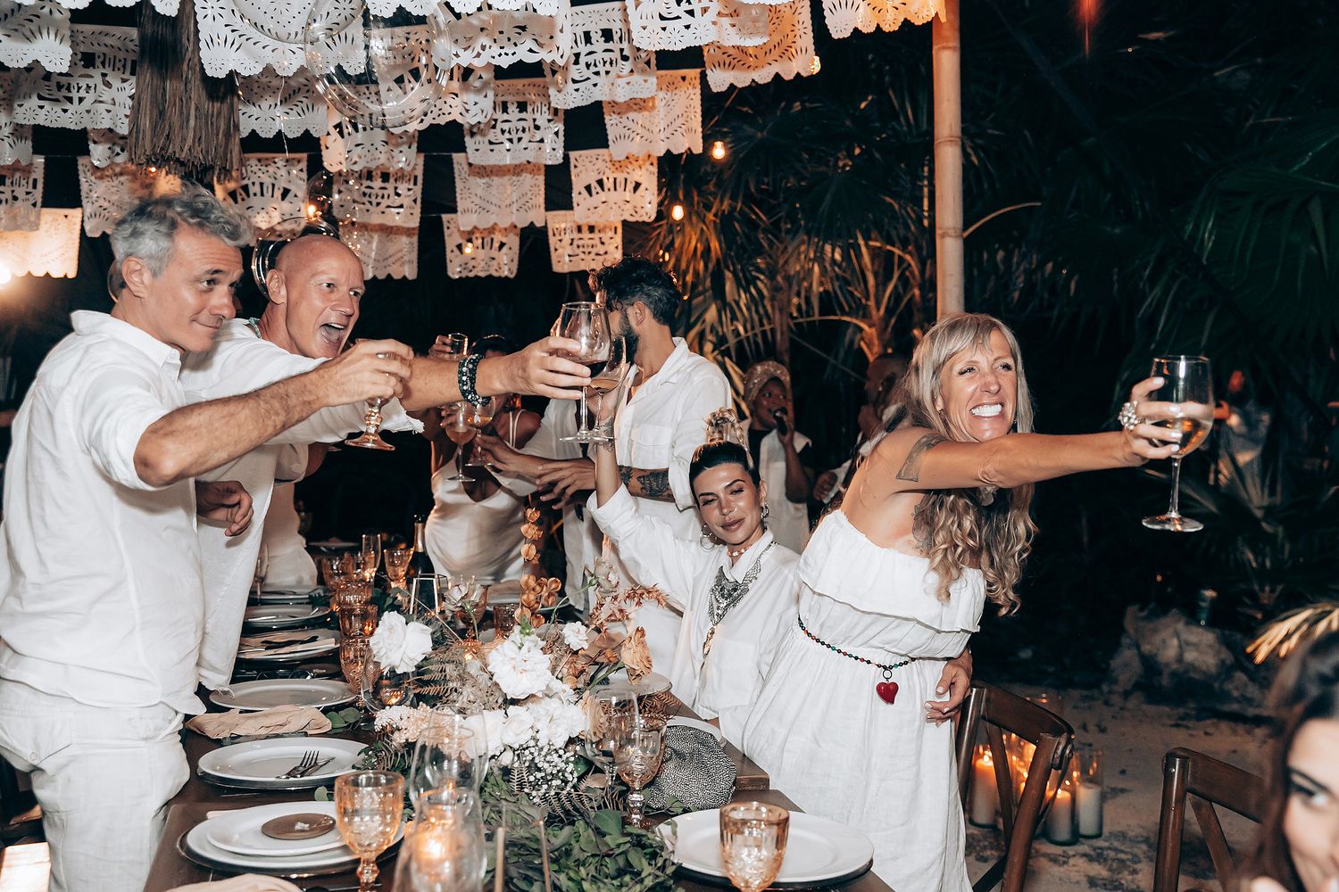 A group of people dressed in white celebrating at a dinner party with plates of food and drinks on the table.