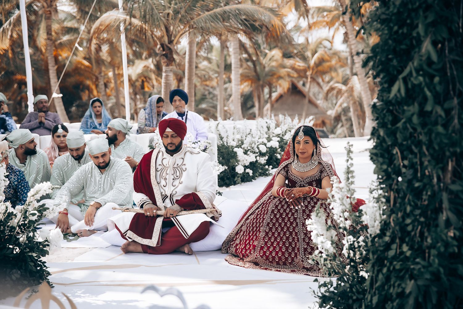 Outdoor wedding setup with white cushions and tropical greenery along a beach.
