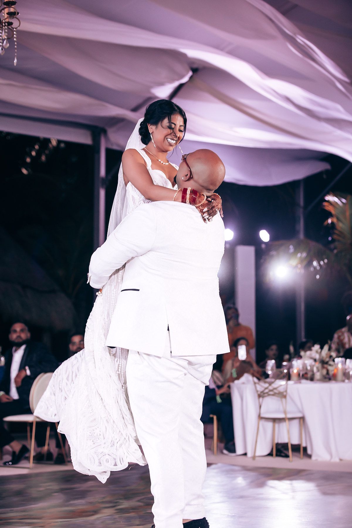 Wedding couple performing romantic dance lifts in matching white attire on illuminated dance floor.