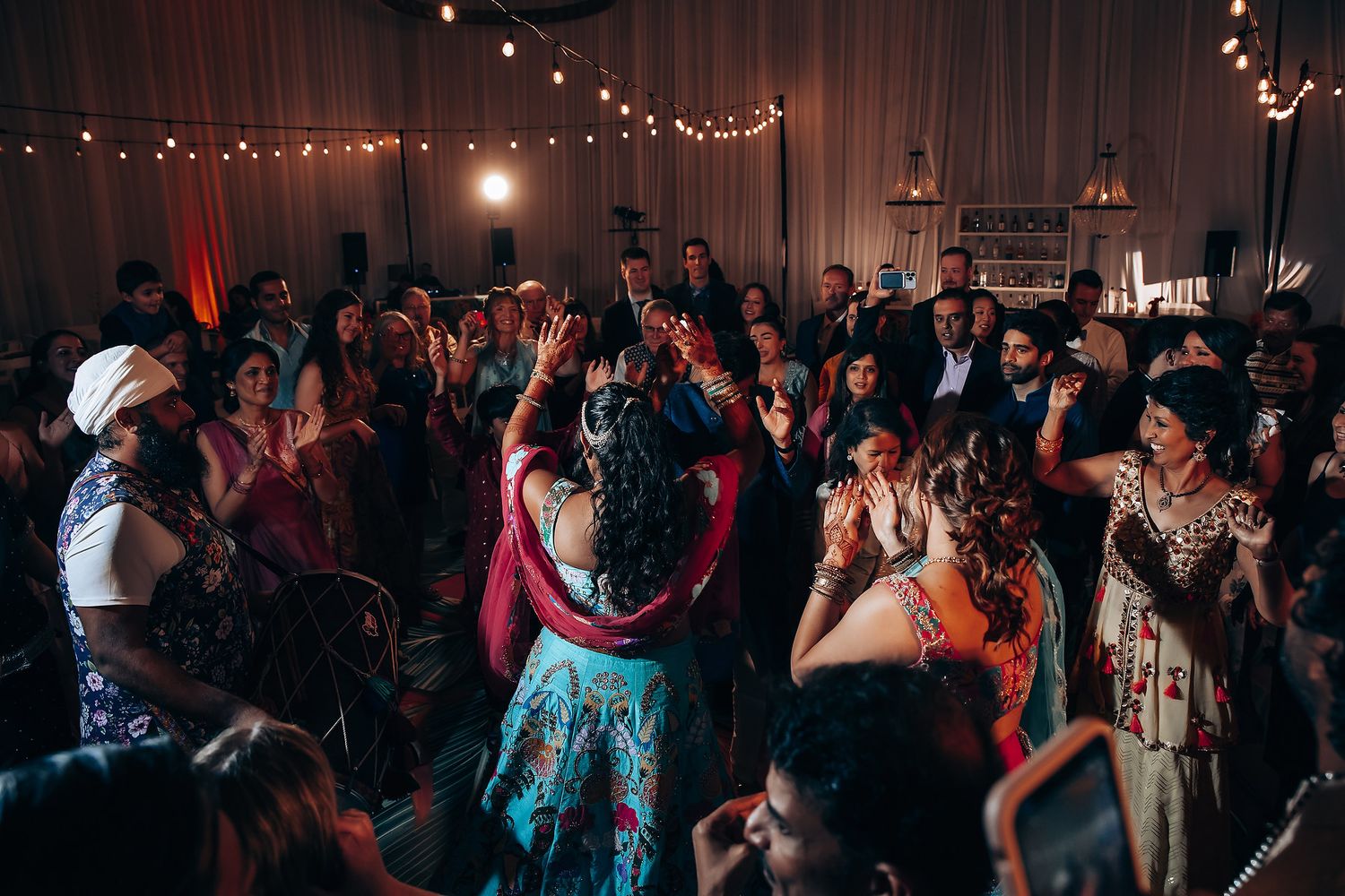 Large crowd dancing and celebrating at a dimly lit indoor reception venue.