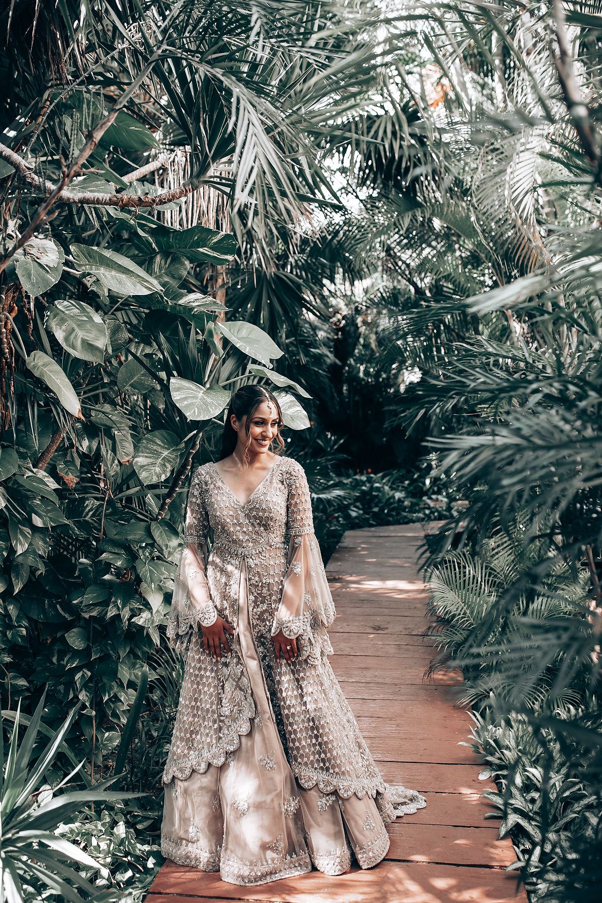 Bride in detailed lace gown walking along wooden pathway through tropical garden setting.