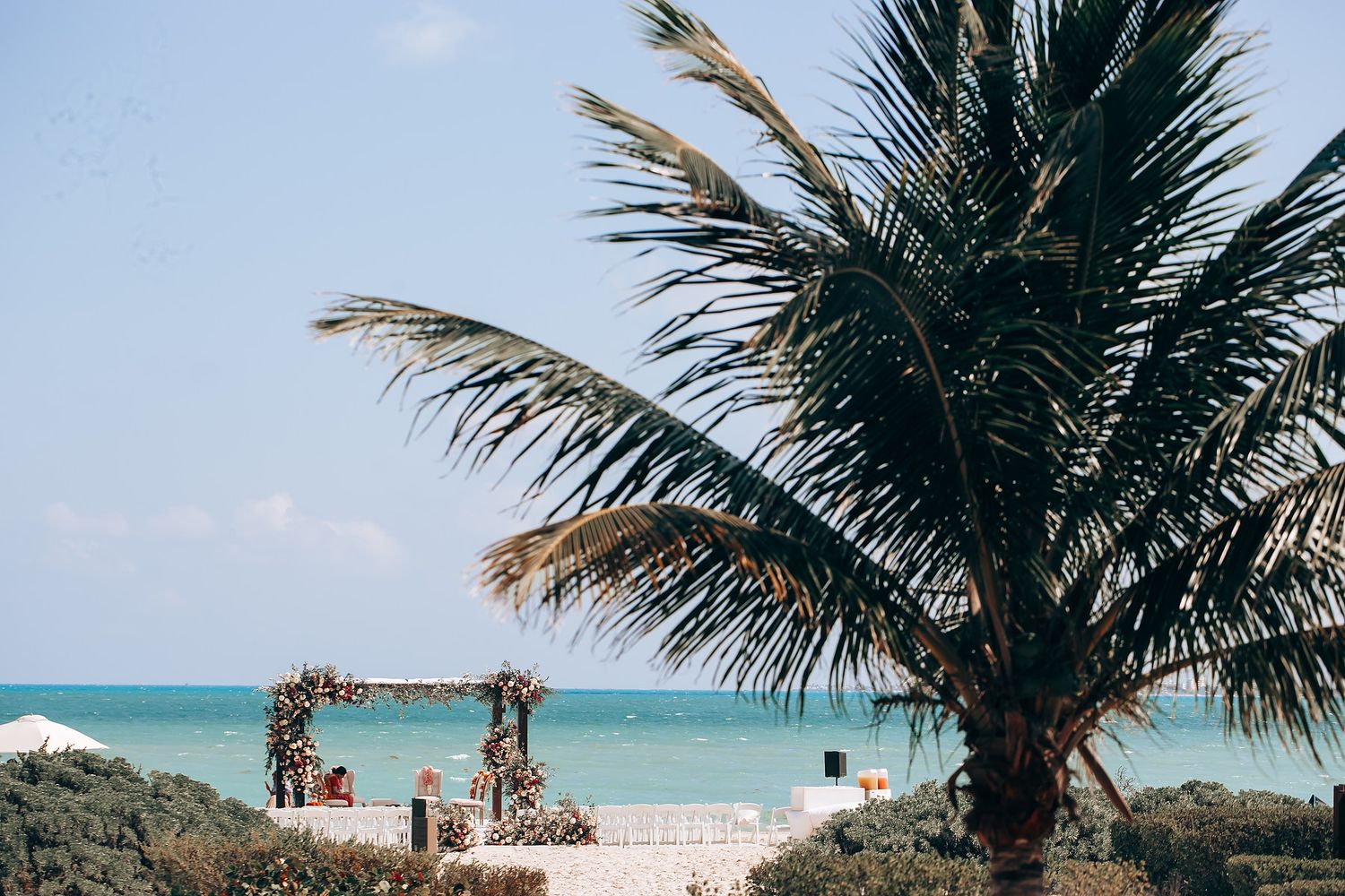 Palm trees frame a beachfront wedding setup with turquoise ocean waters in background.