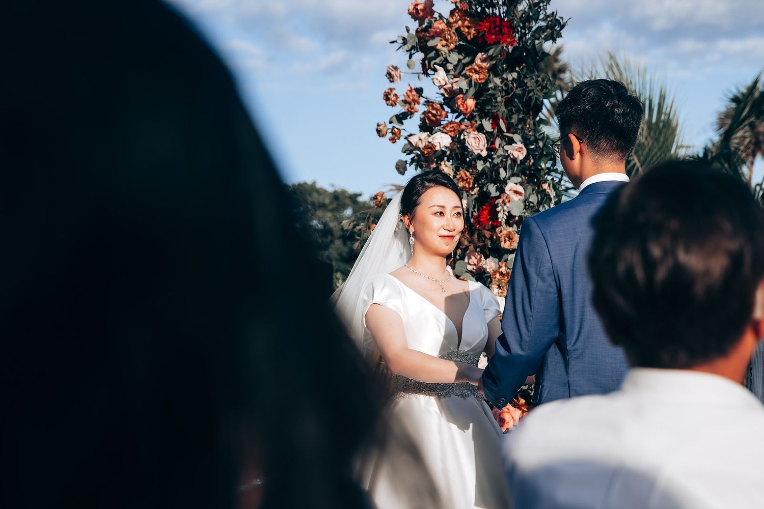 Wedding ceremony under blue skies with floral arch and gathered guests.