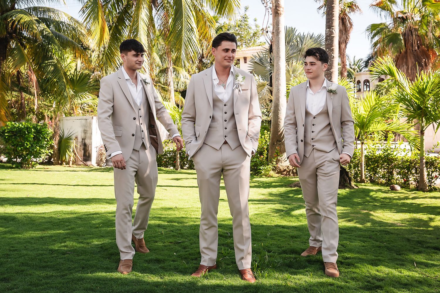 Groomsmen in light gray suits pose together on a sunny lawn with palm trees in the background.