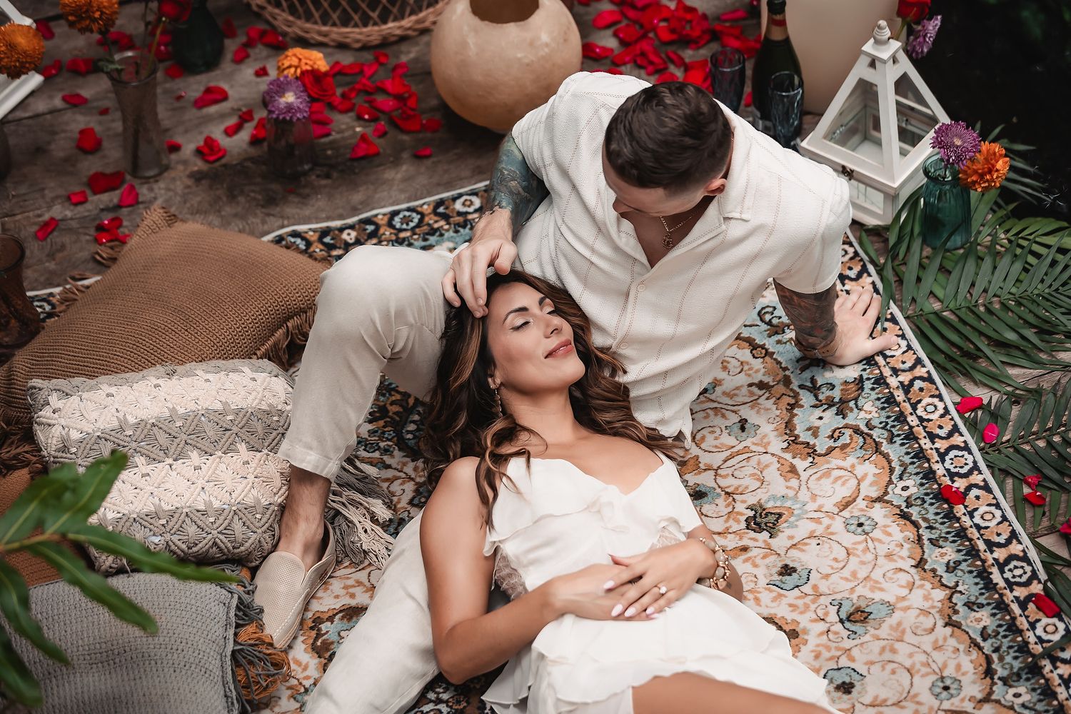 Couple in white outfits sharing an intimate moment on a decorated outdoor rug with rose petals.