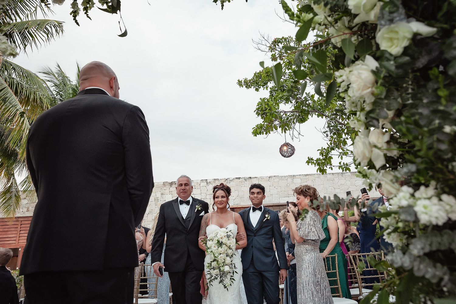A wedding party gathers outdoors under palm trees and floral decorations during a sunny celebration.