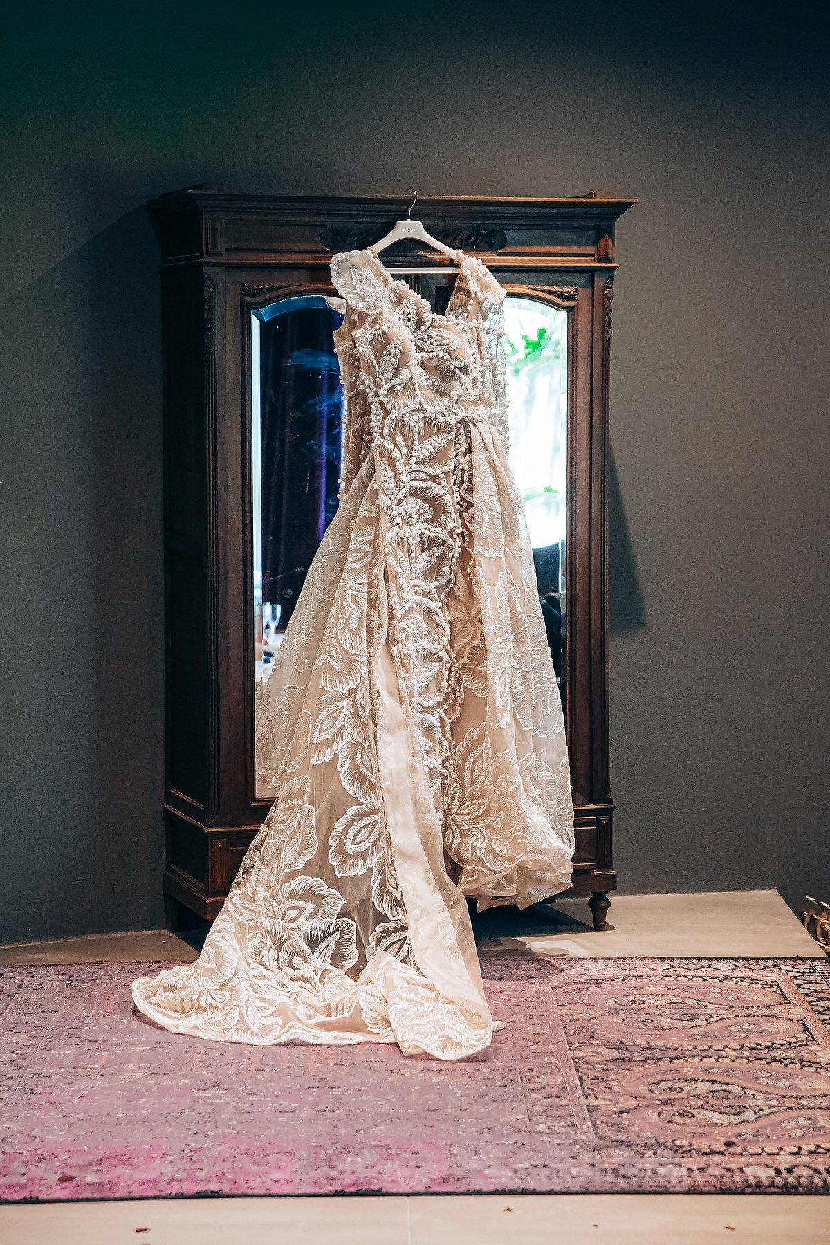 A delicate lace wedding dress with intricate embroidery details hangs elegantly against a dark wall with pink carpet below.