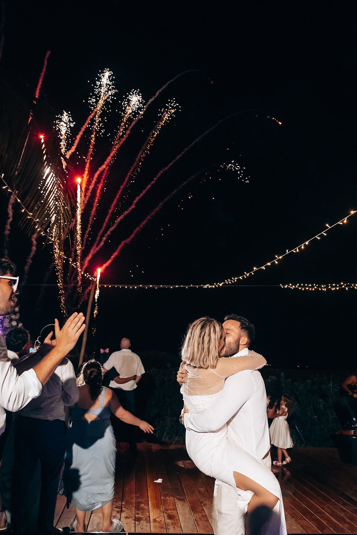 Wedding party dances under fireworks display with string lights creating magical nighttime ambiance.