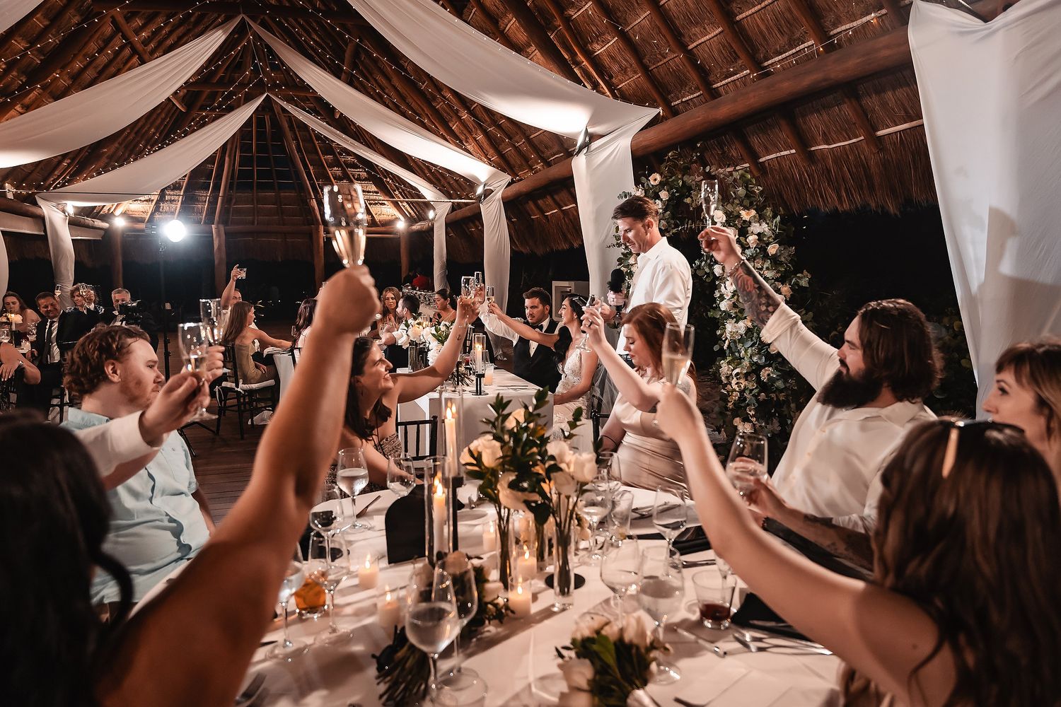 Wedding reception guests raise their glasses in celebration under rustic wooden beams.