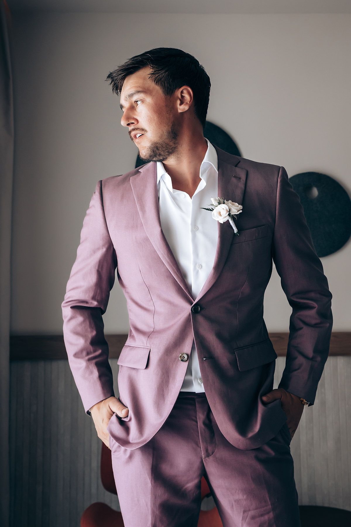 A man in a dusty pink suit jacket and white shirt poses against a wall in a series of fashion photographs.