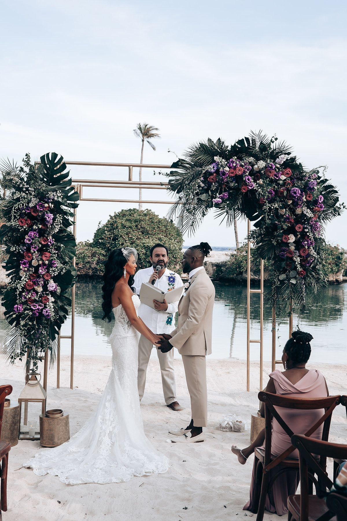 Wedding ceremony under a floral arch with pink blooms against a waterfront backdrop.