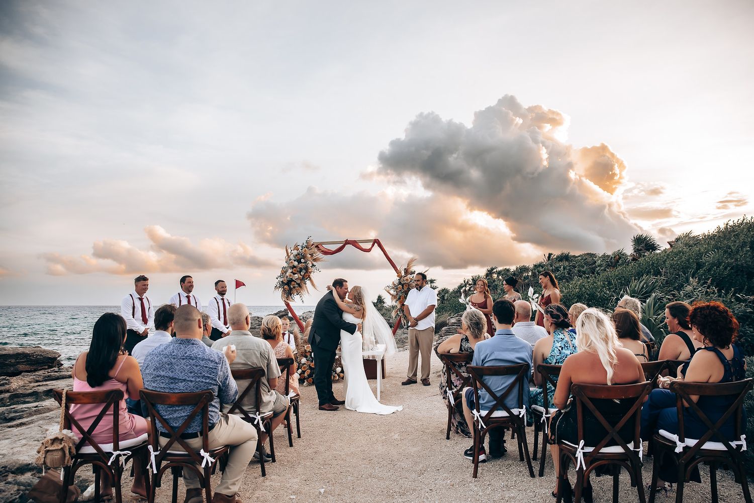 A panoramic view of an outdoor beach wedding ceremony with guests seated in white chairs at sunset.