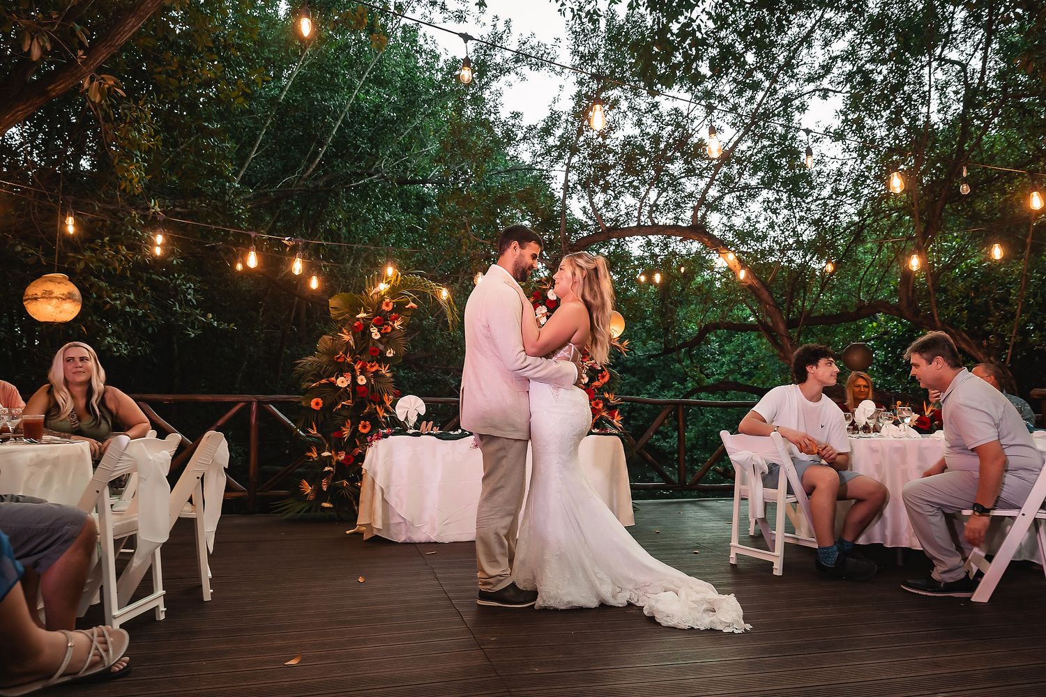Couple shares first dance on wooden deck under twinkling lights at outdoor reception.
