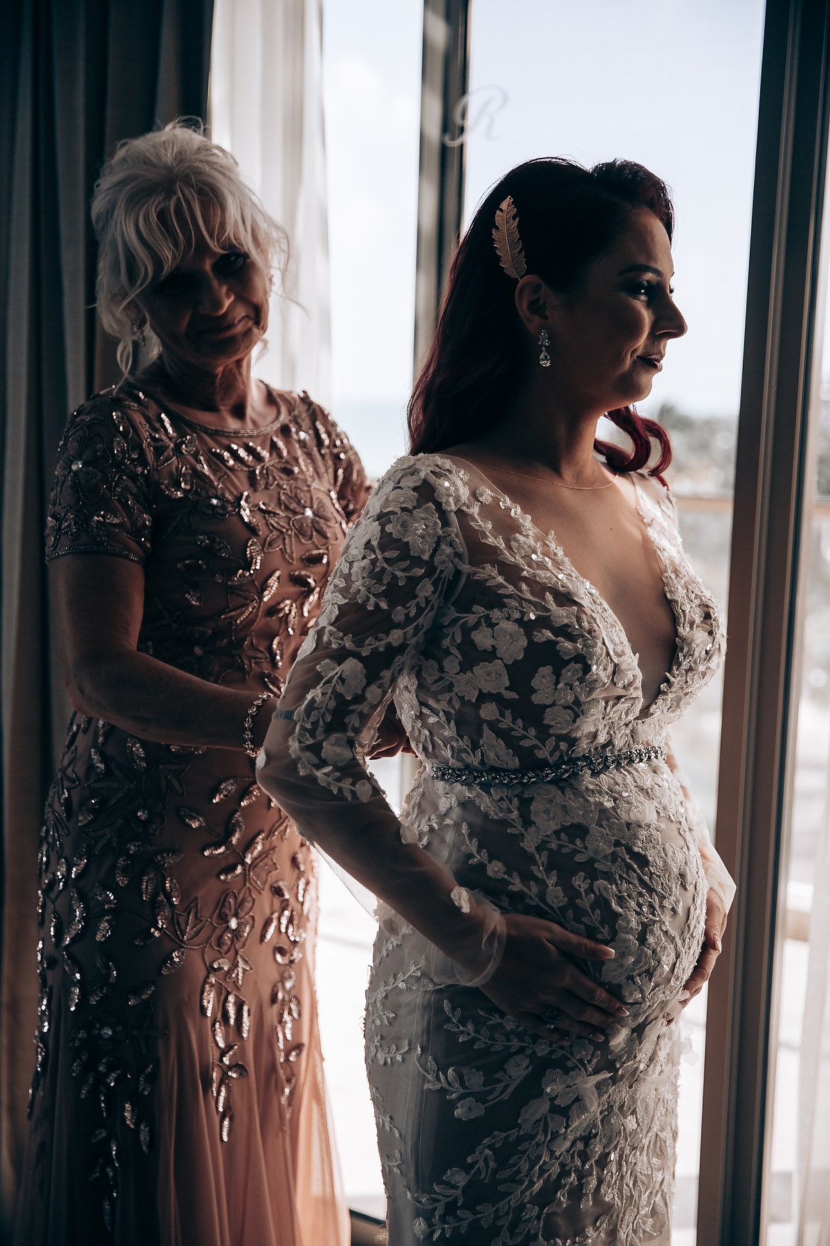 A group of women in elegant evening gowns stand together by a window in soft lighting.